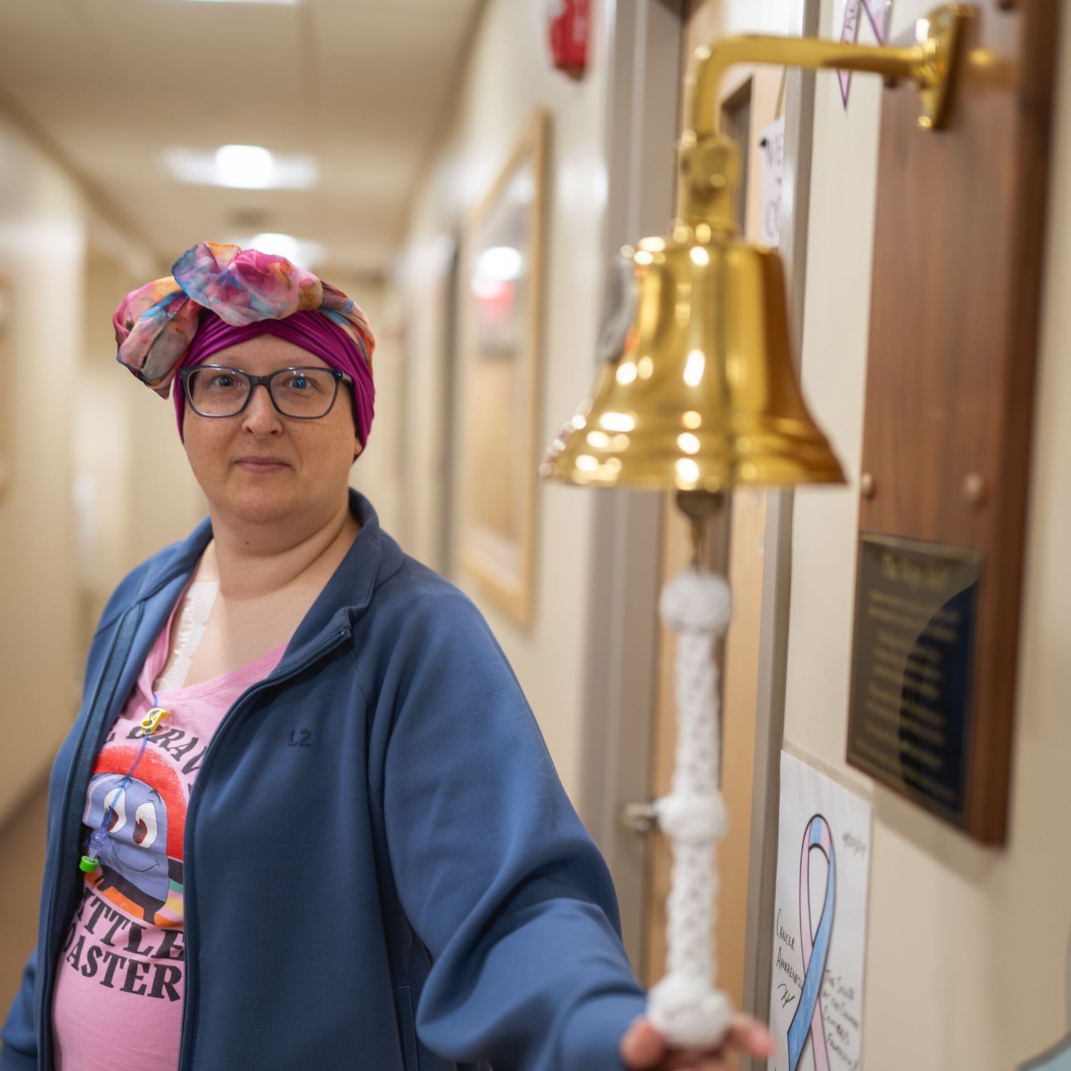 Woman in pink headscarf and glasses stands beside a brass bell in a hospital hallway, symbolizing cancer treatment completion.