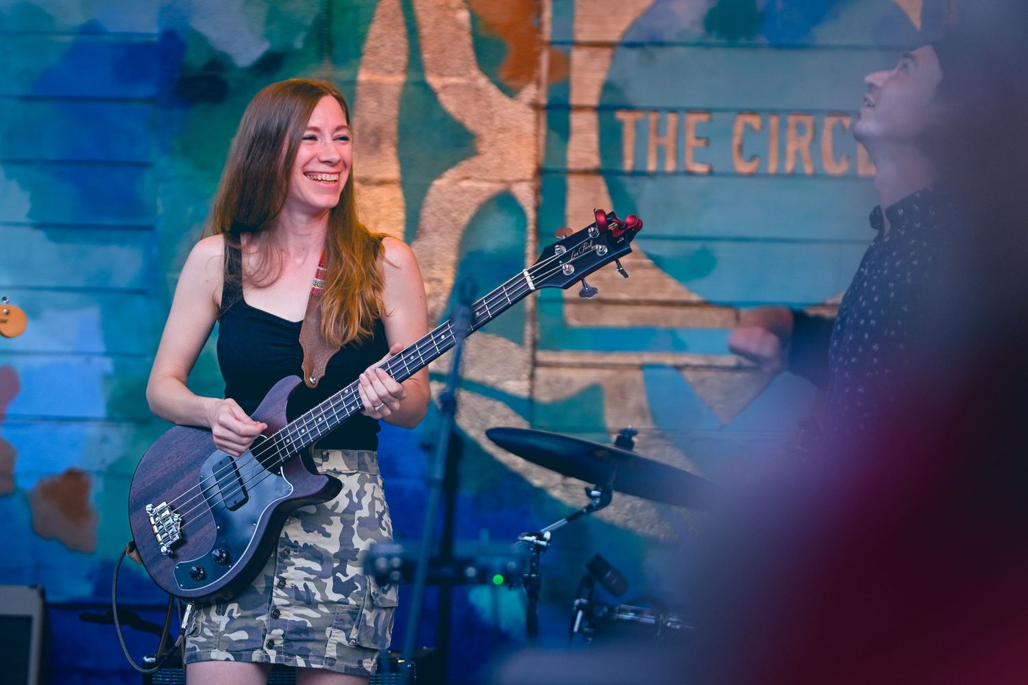 The Mimic band: A musician performs on stage with a bass guitar in front of a colorful mural with blue tones and text.