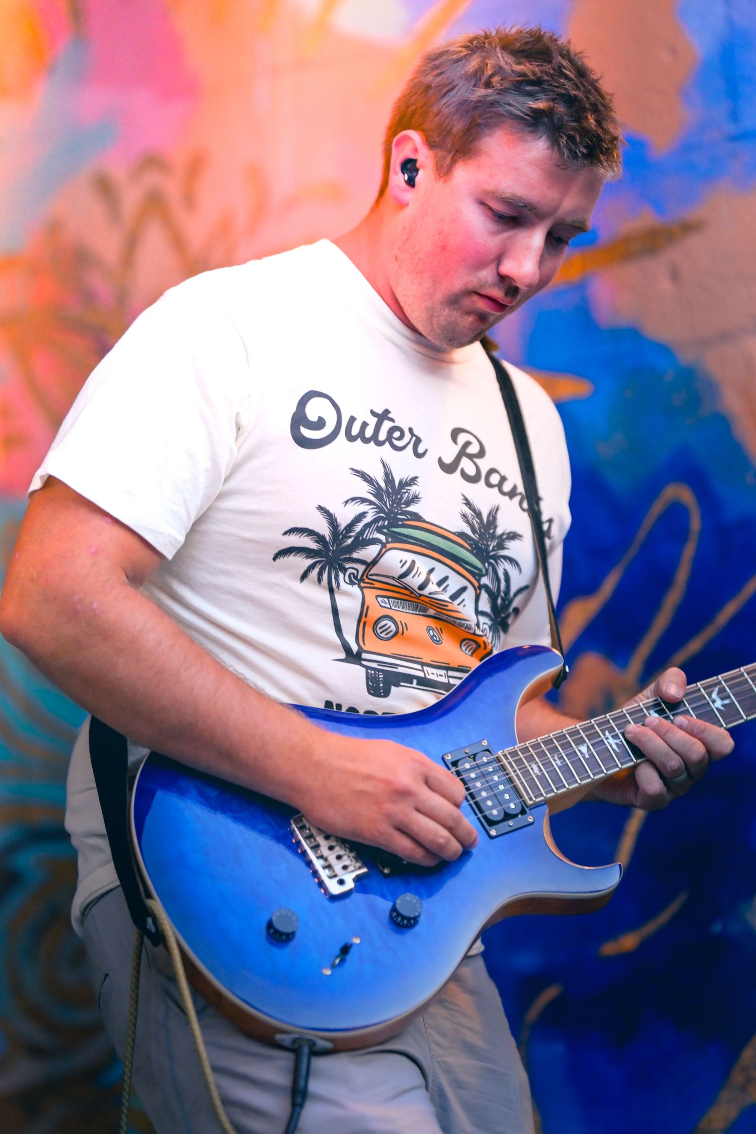 Musician in Outer Banks t-shirt plays blue electric guitar against colorful graffiti wall background.