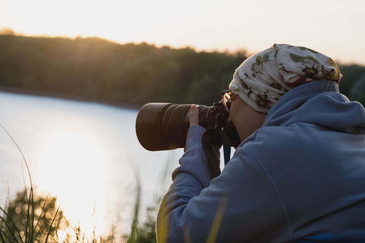 Photographer with telephoto lens capturing sunset over a lake, wearing a camo hat and gray hoodie outdoors.