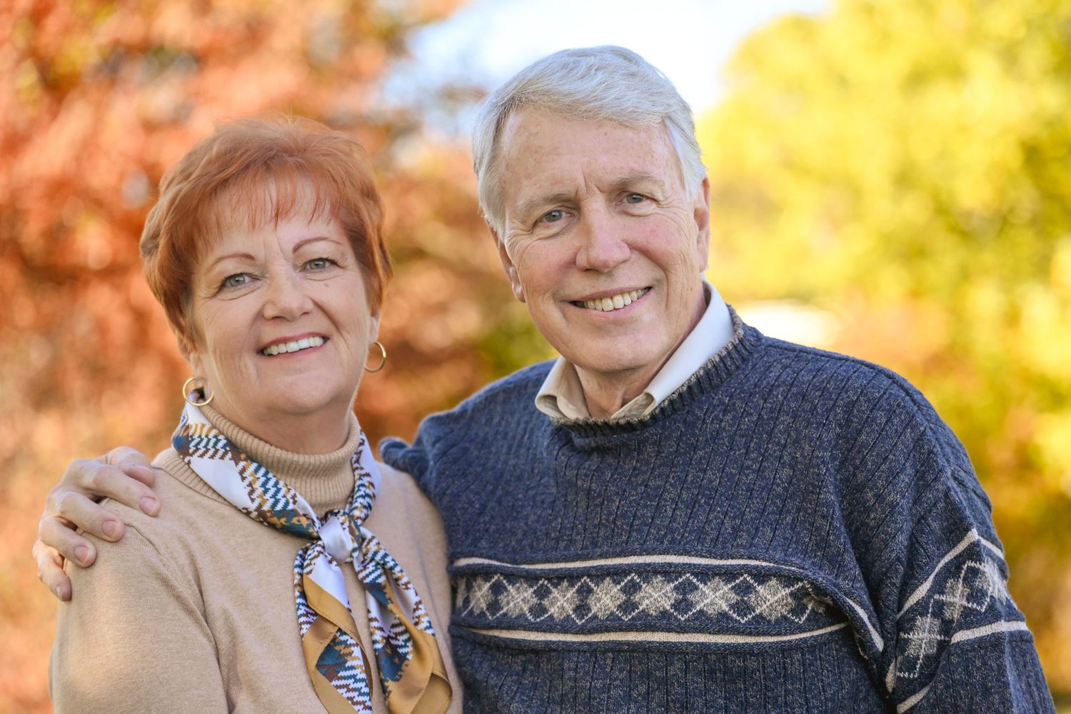Smiling senior couple posing outdoors in autumn, man in blue sweater with arm around woman in colorful scarf.