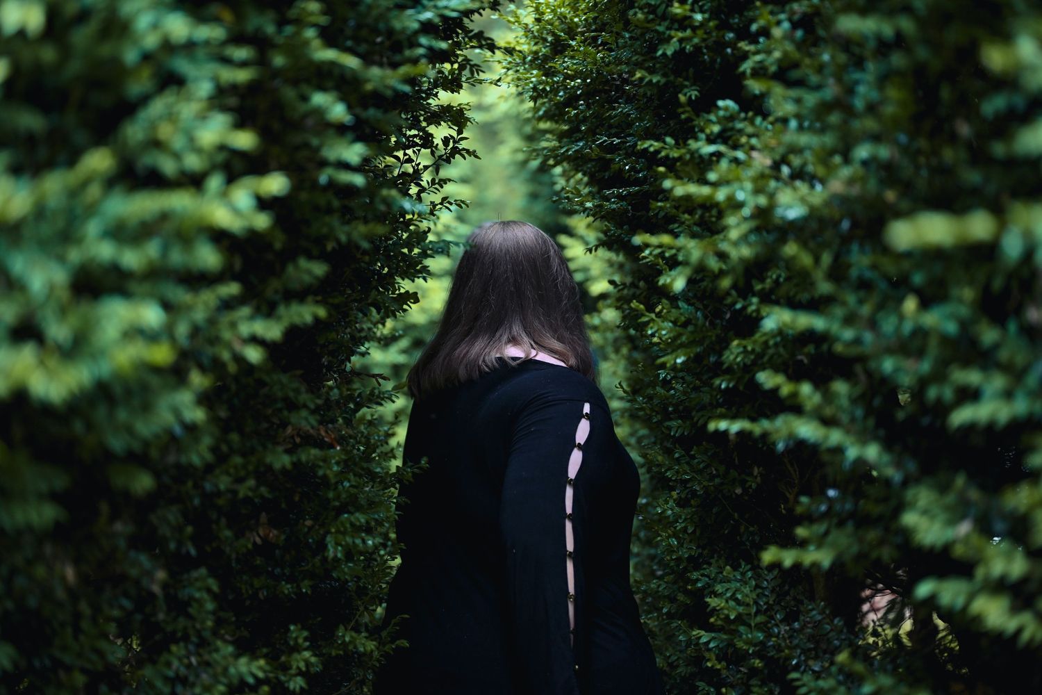 A figure in a dark coat stands between lush green hedges on a garden path.