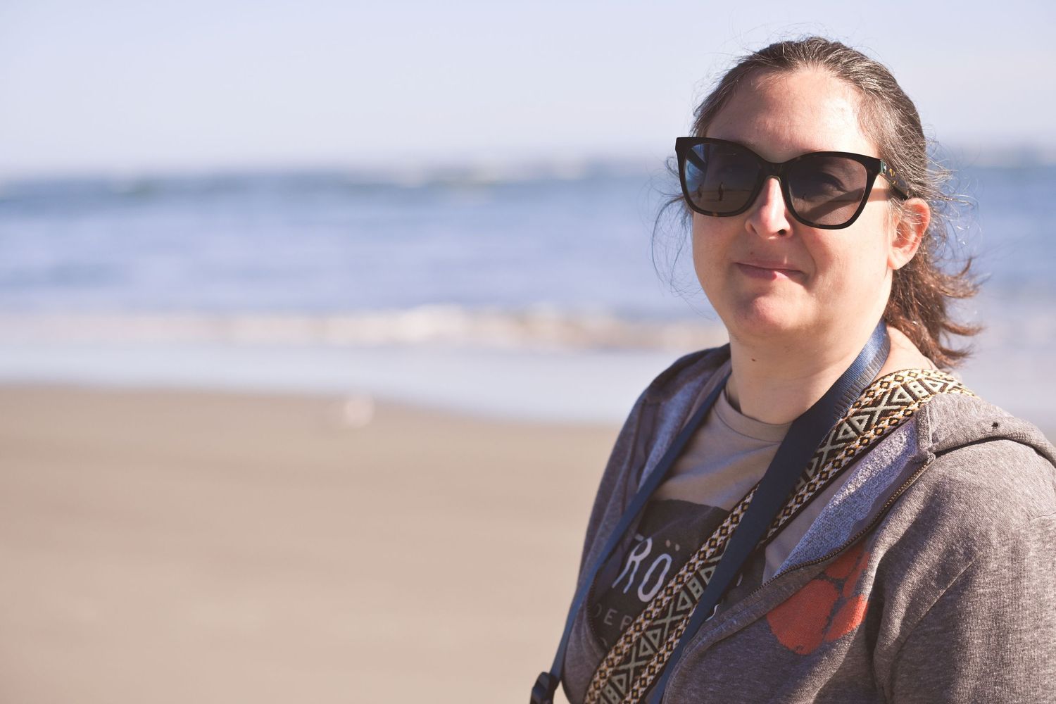 A person wearing sunglasses and a scarf walks along a sunny beach on a breezy day.