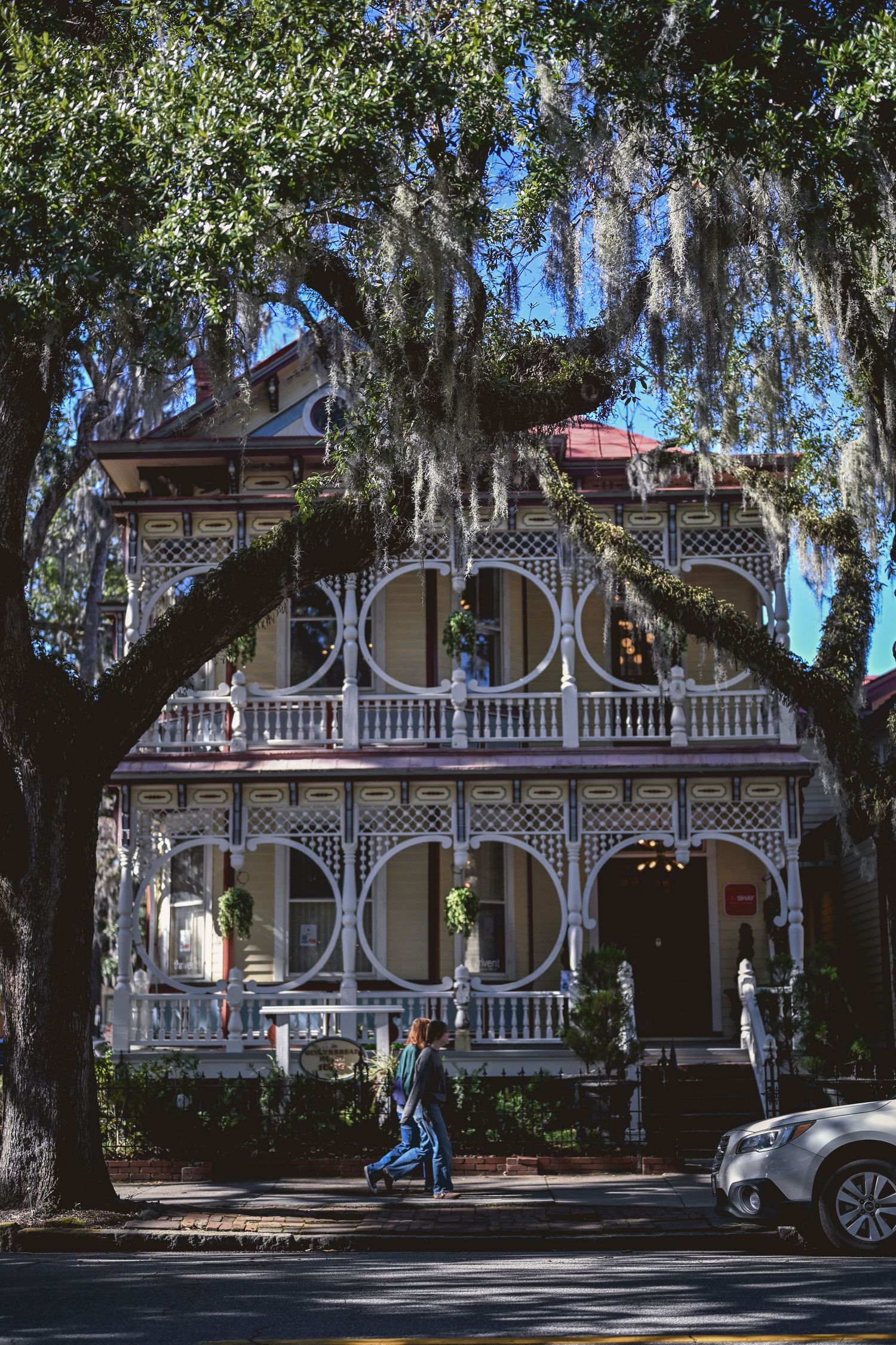 Historic Victorian mansion with ornate white lattice porches and Spanish moss draped oak trees in the foreground.