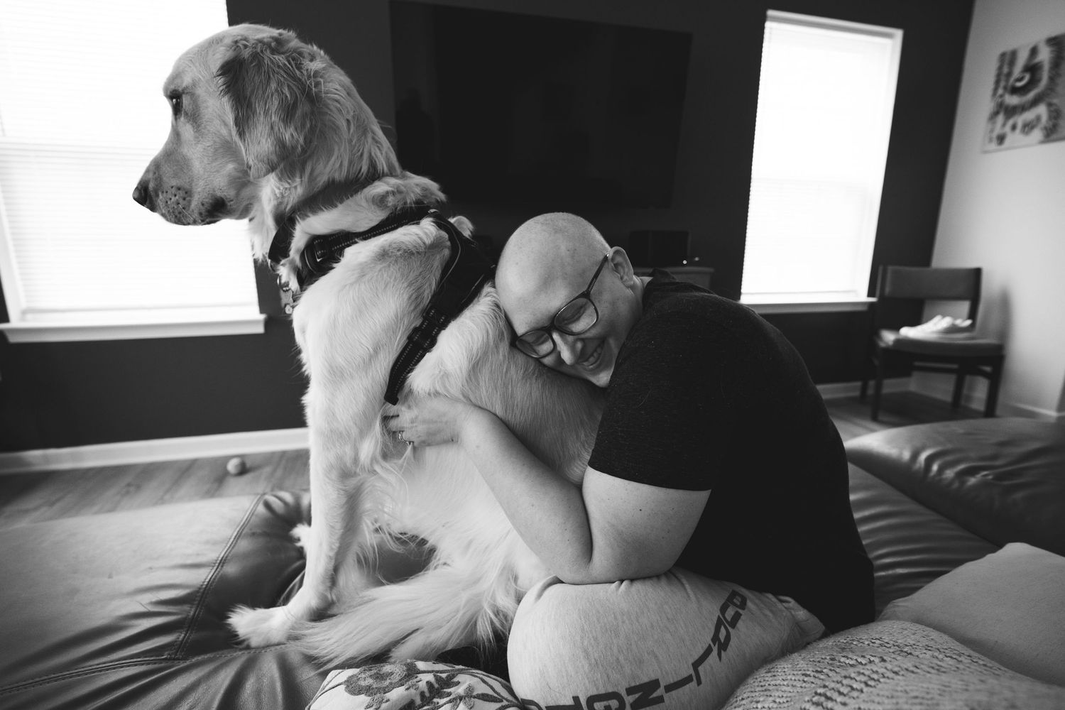 Bald man hugging a large dog on a bed in a black and white indoor photo.