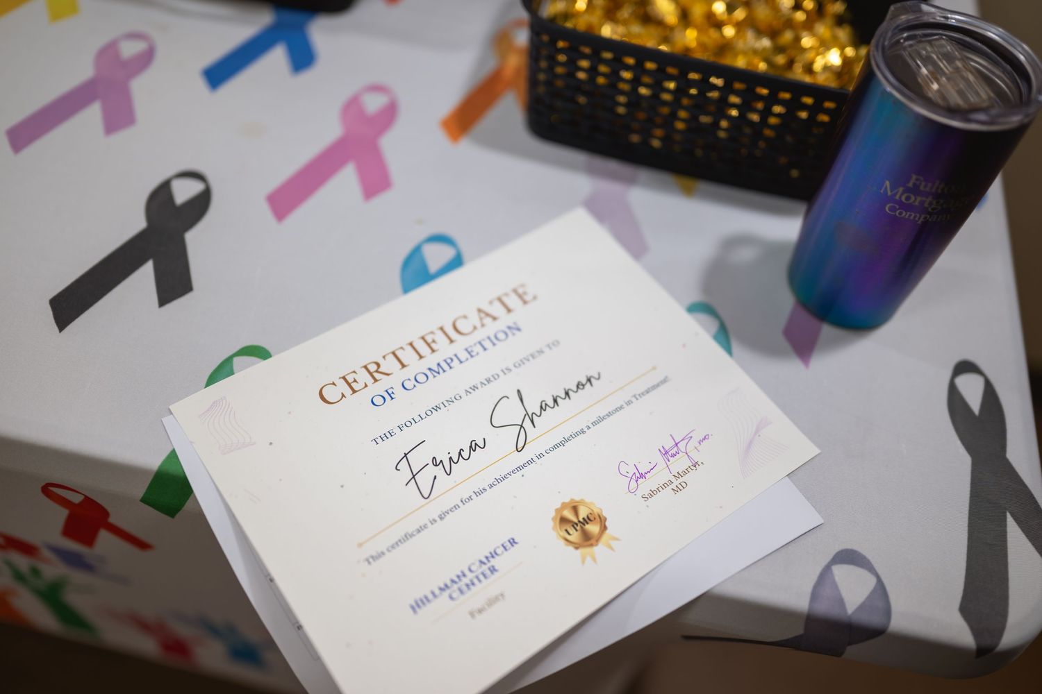 Certificate of Completion for Erica Shannon displayed with colorful cancer awareness ribbons, a tumbler, and gold basket.