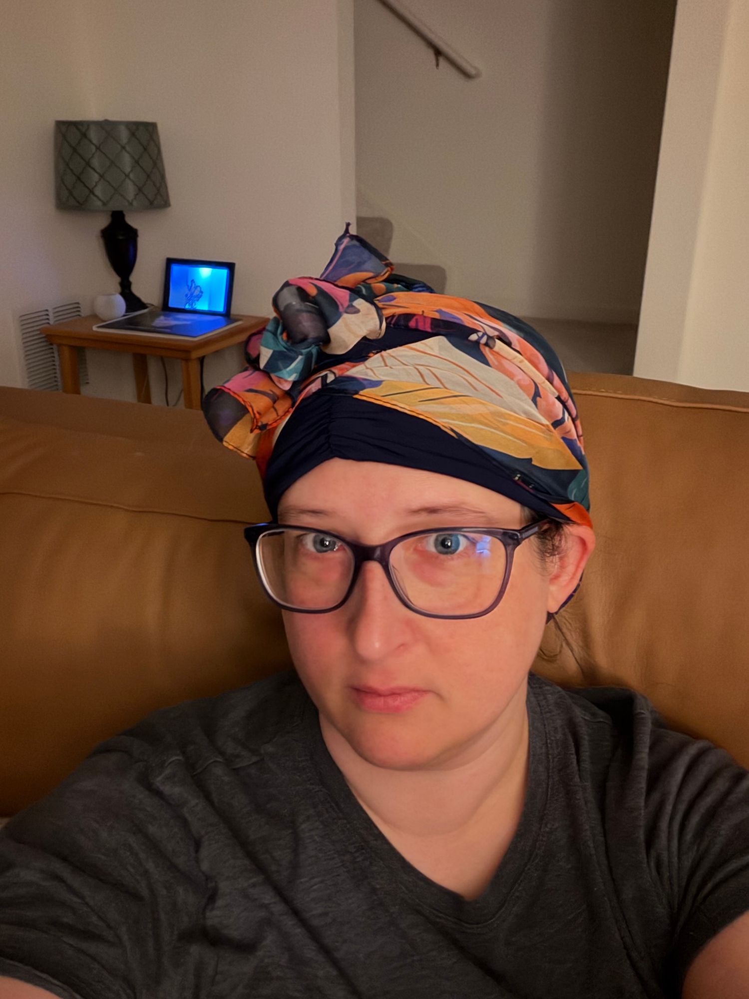 Woman wearing colorful patterned head scarf and glasses sitting on brown leather sofa indoors.