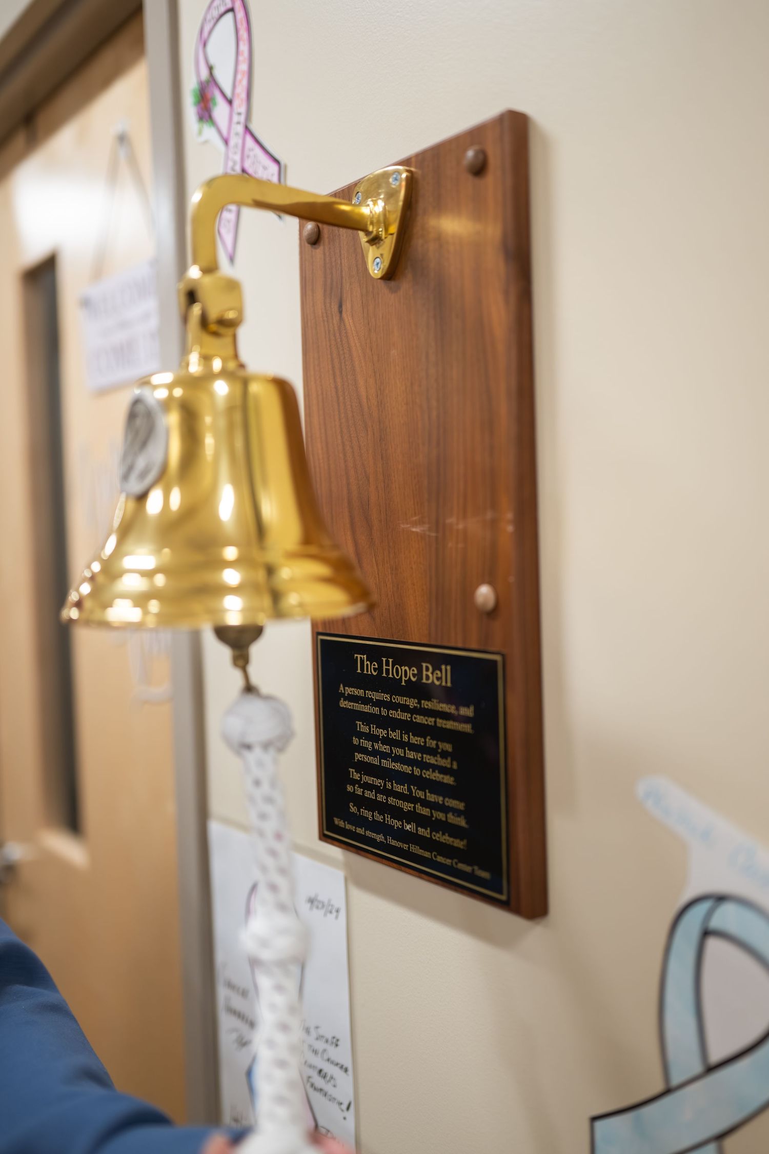 Gold Hope Bell mounted on wooden plaque in hospital hallway, symbolizing cancer treatment completion.