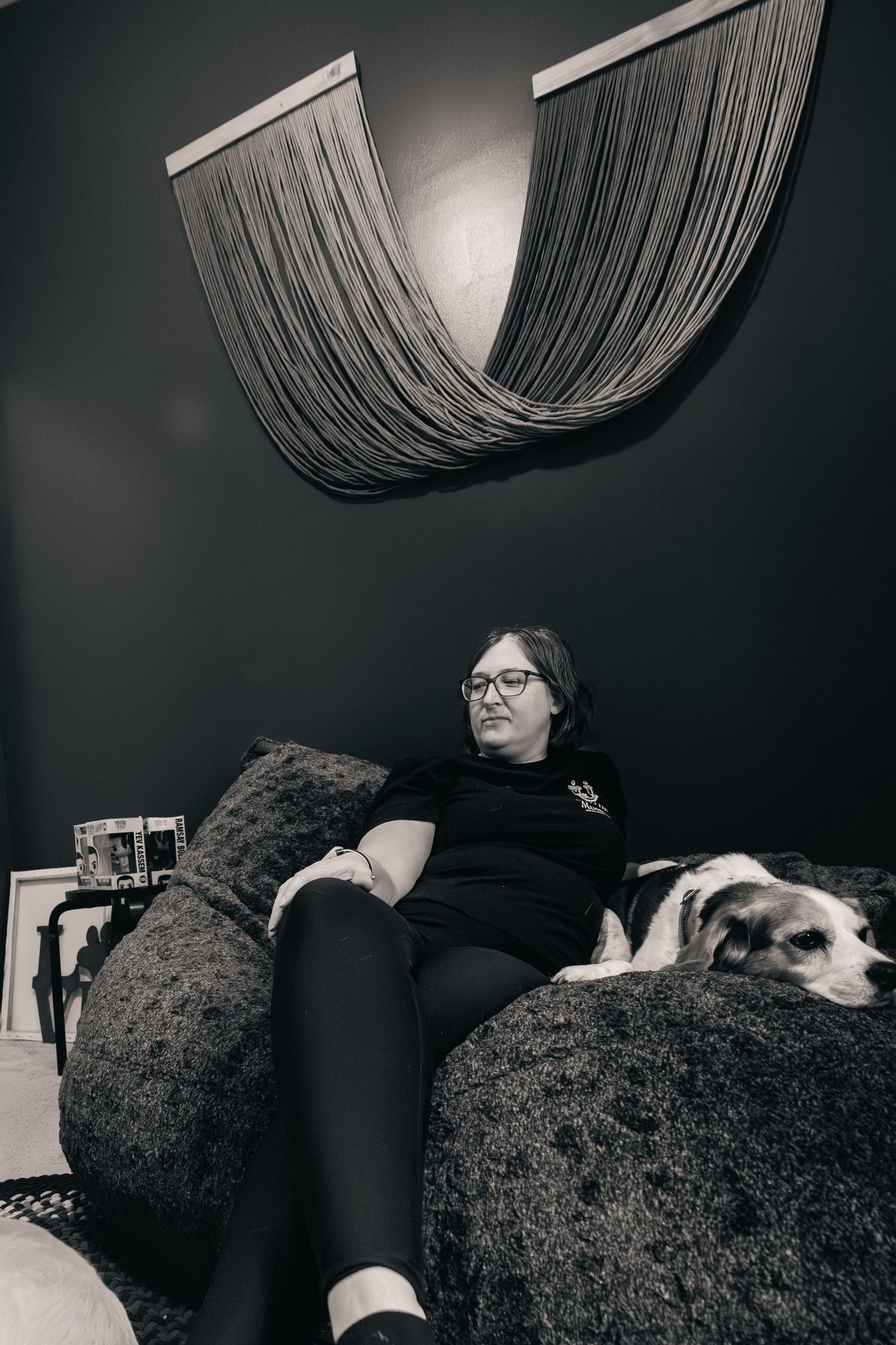 Woman with glasses relaxing on large bean bag chair with a dog in a dark moody room, black and white photo.