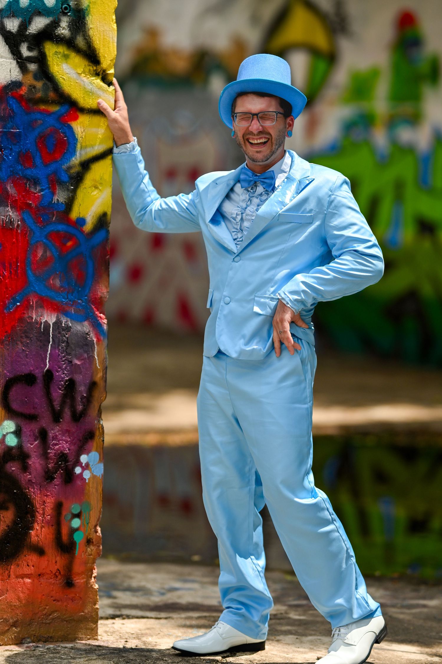 Dancer in light blue jumpsuit performs dynamic dance moves against colorful graffiti wall background.