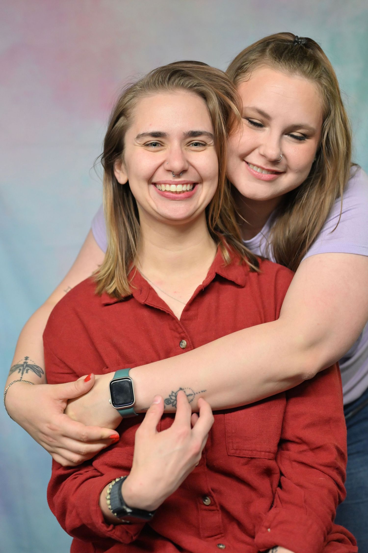 Two people embrace in a candid studio portrait against a mottled blue background, wearing red and purple tops.