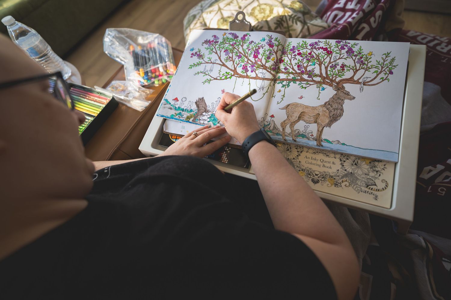 Person coloring an intricate adult coloring book page featuring a deer and tree with colored pencils on a table.