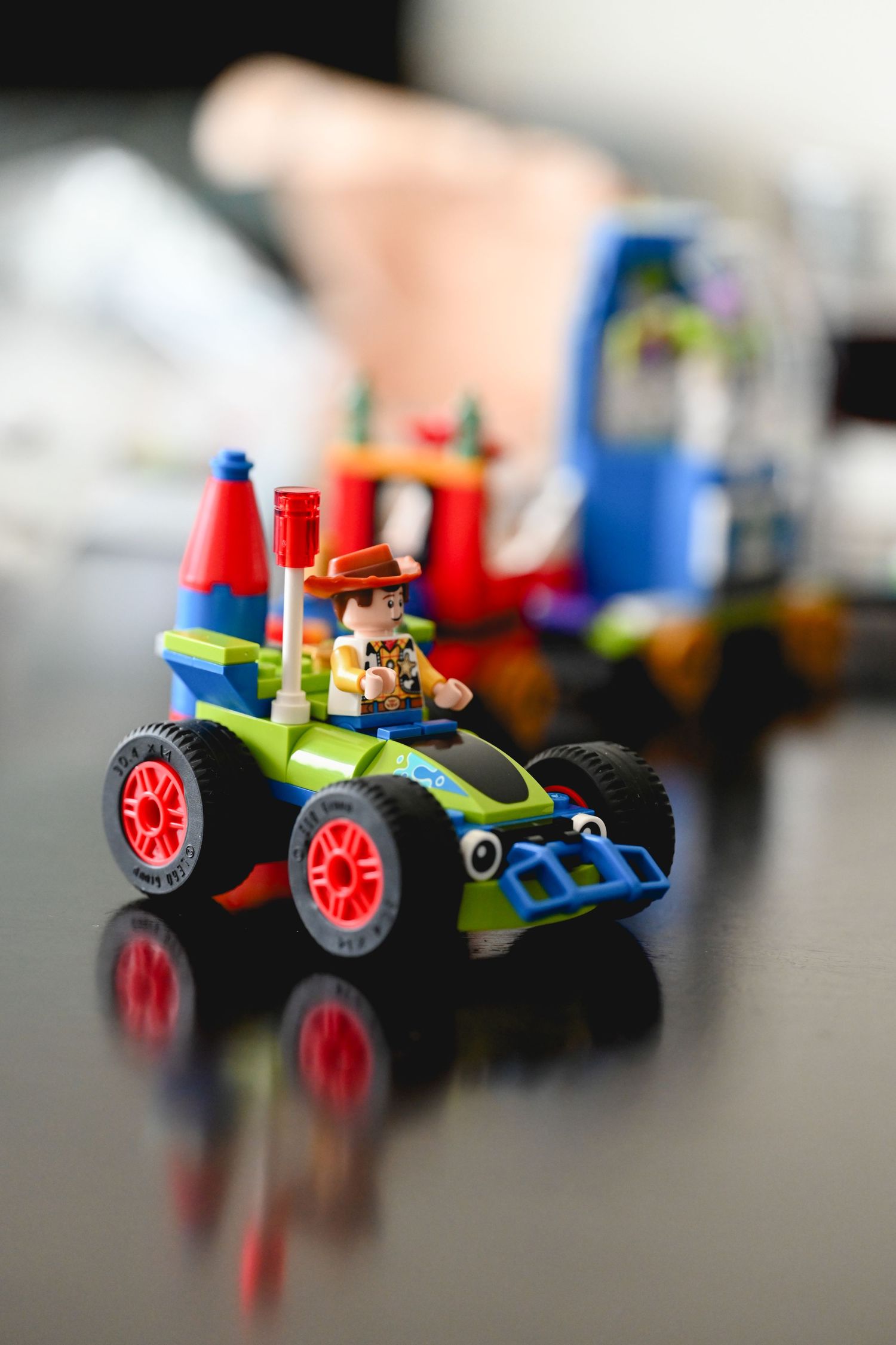 LEGO Toy Story Woody minifigure riding RC car toy on reflective black surface with blurred colorful LEGO set background.