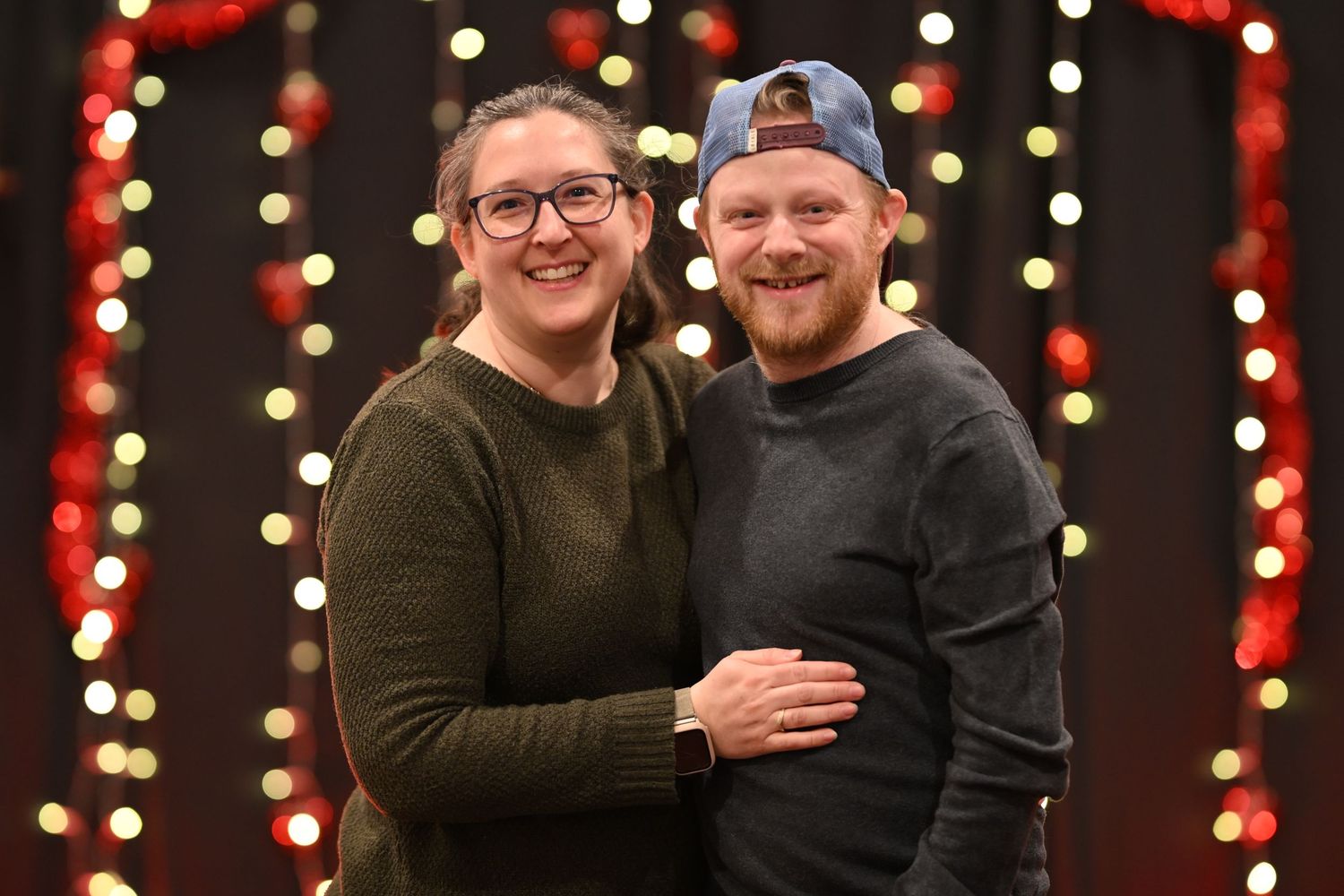A couple poses together in front of a festive holiday backdrop with red lights and bokeh effects.