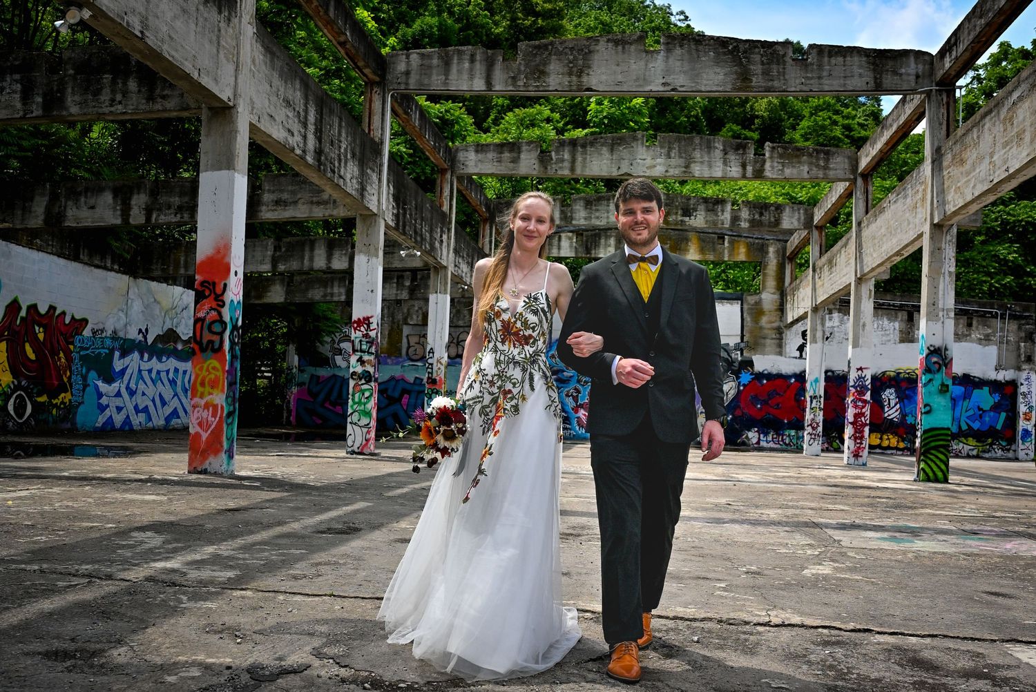 A couple in formal attire poses together in an abandoned urban space with graffiti-covered walls and concrete pillars.