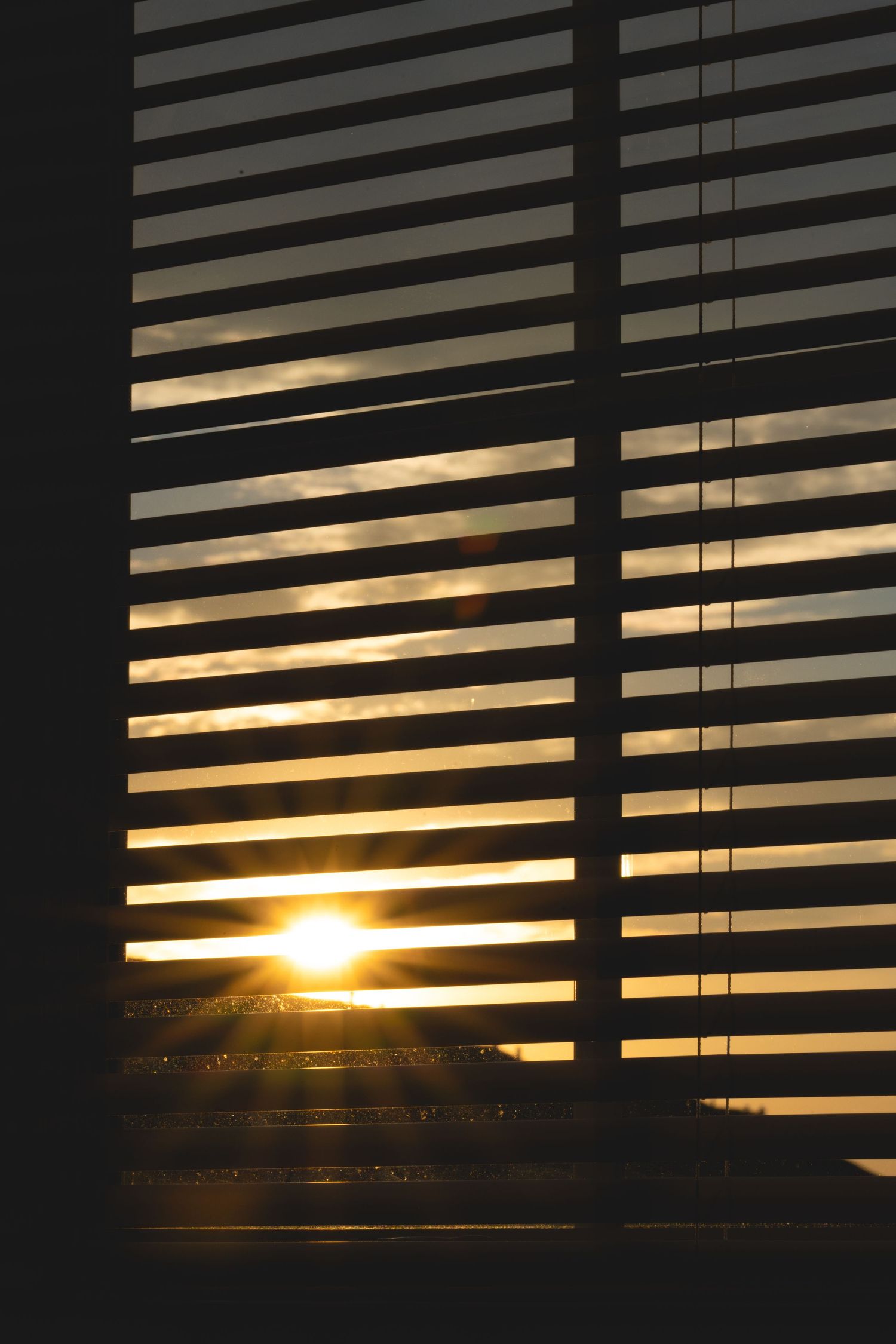 Warm golden sunset viewed through horizontal window blinds casting dramatic striped shadows in a darkened room.
