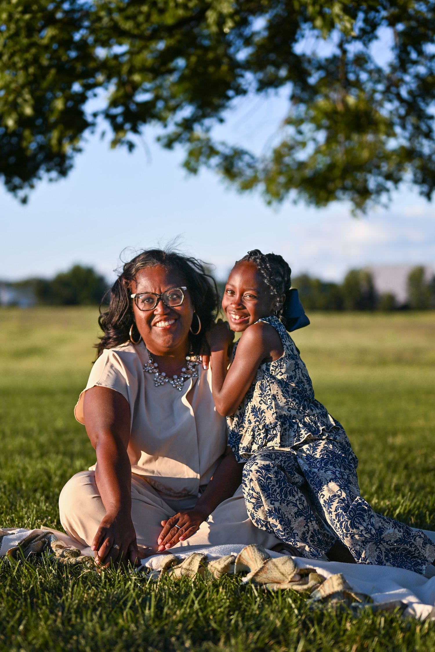 A woman and young girl smile together on a blanket in a sunny park, framed by green tree branches.
