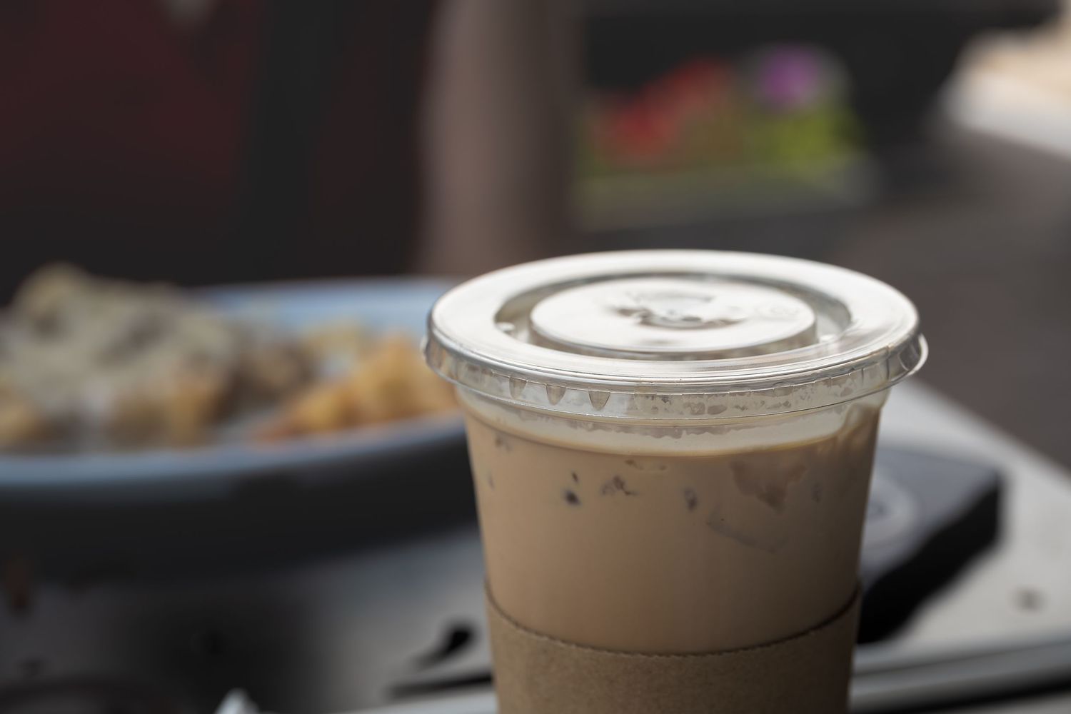 Iced coffee drink in a clear plastic cup with lid on an outdoor table with blurred food in background.