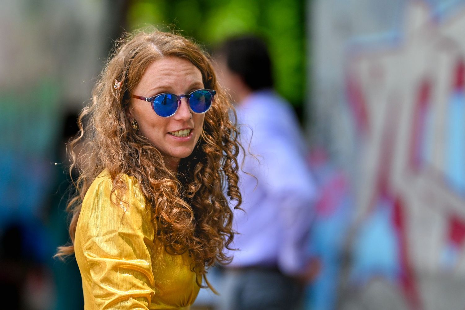 Person wearing yellow jacket and blue sunglasses with long curly hair smiling on blurred urban background.