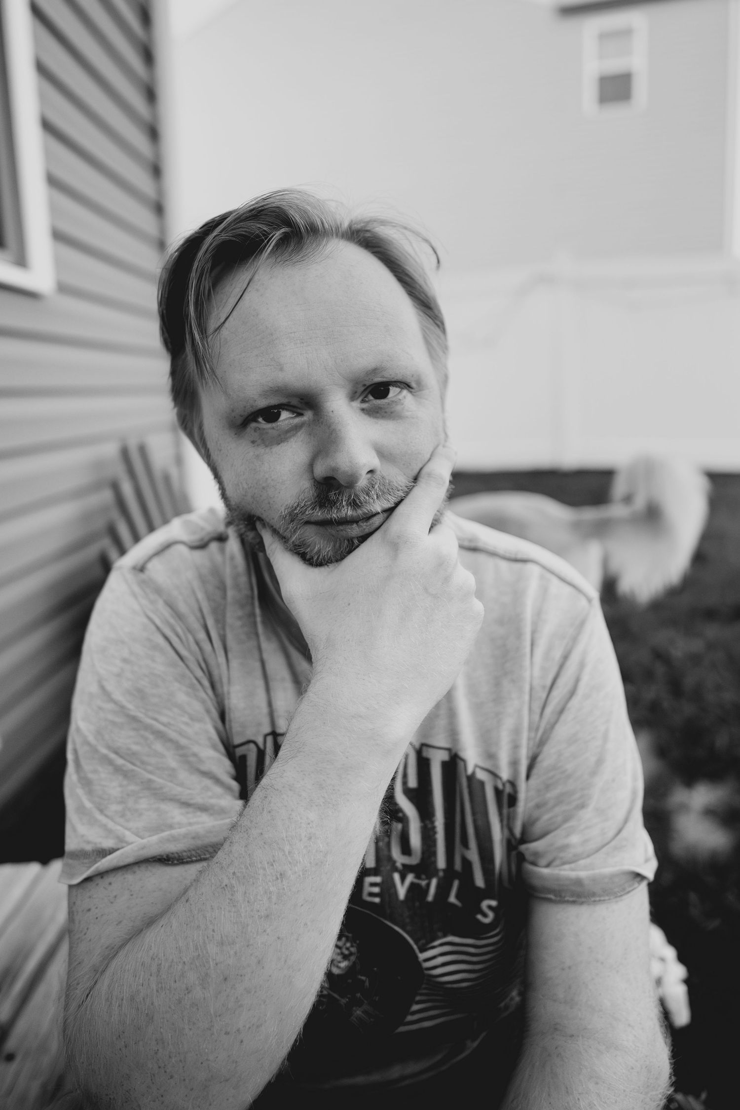 Black and white portrait of a thoughtful man with beard resting chin on hand, seated outdoors near a house.