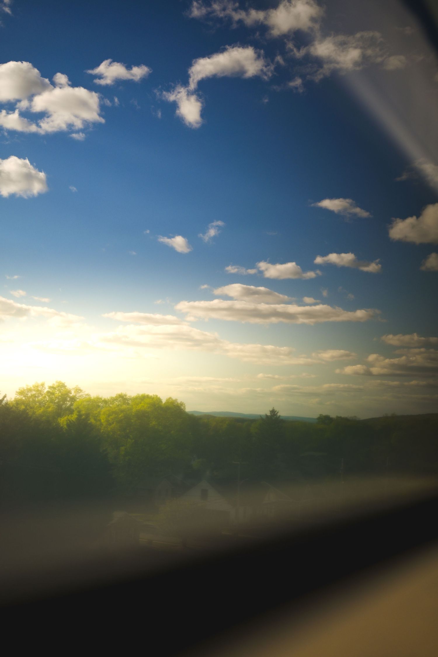 Scenic view through a car window of a golden sunset over lush green trees under a blue sky with white clouds.