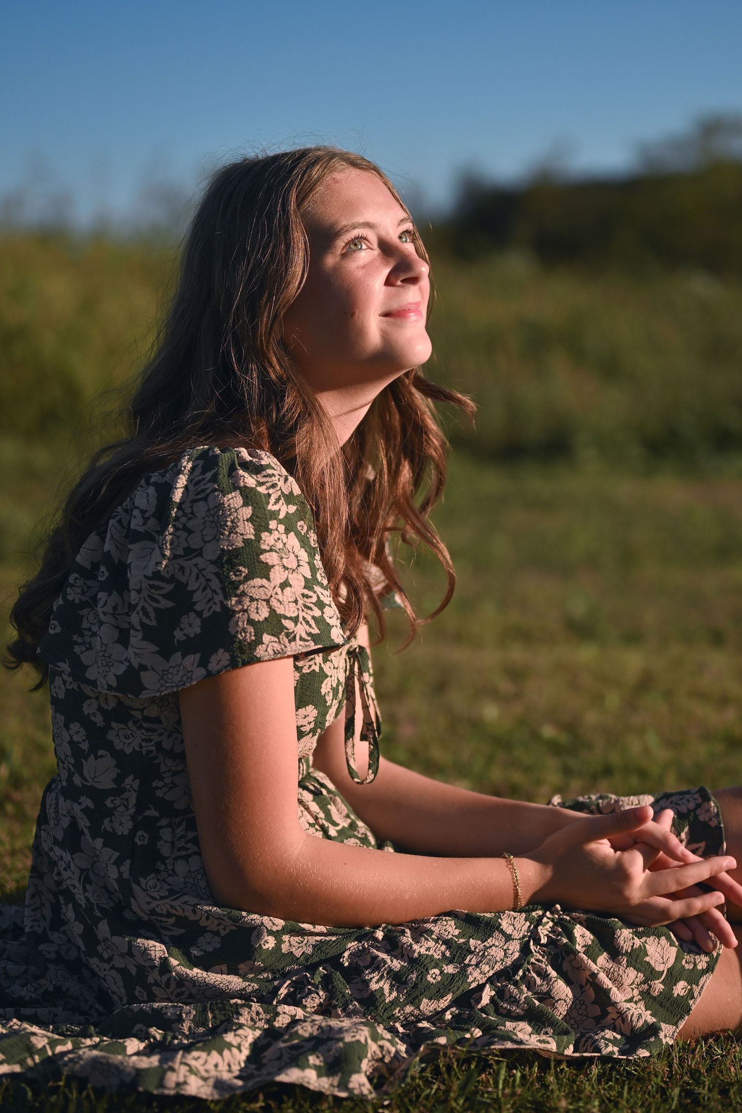 A young person in a floral dress sits in a grassy field during golden hour, looking upward at the sky.