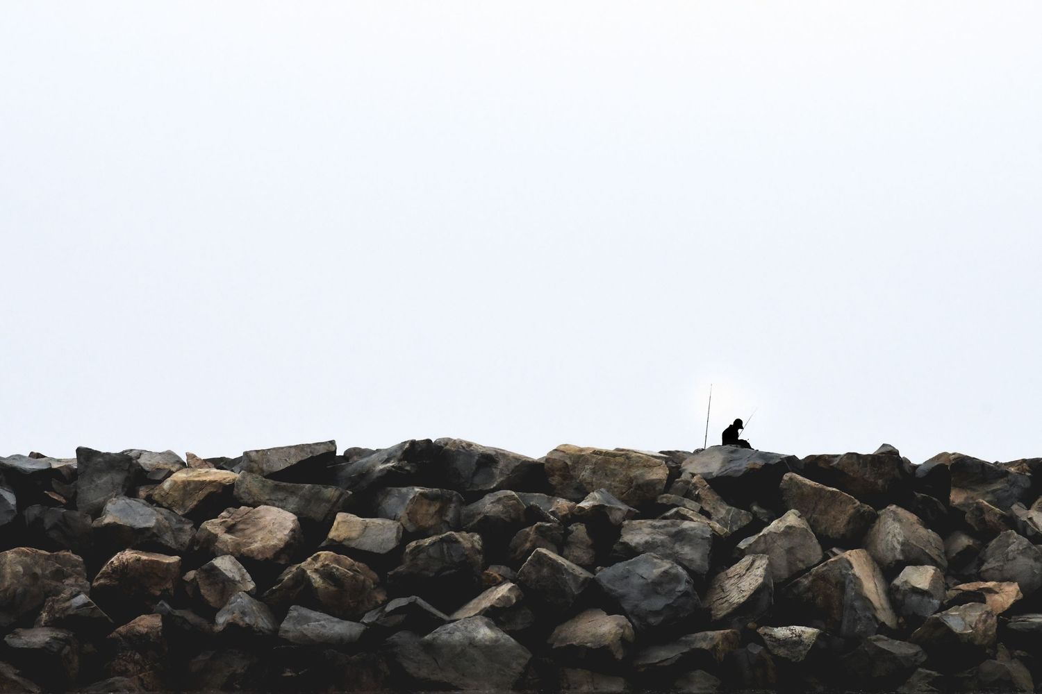 A lone silhouette sits atop a rugged rocky breakwater against a pale overcast sky.