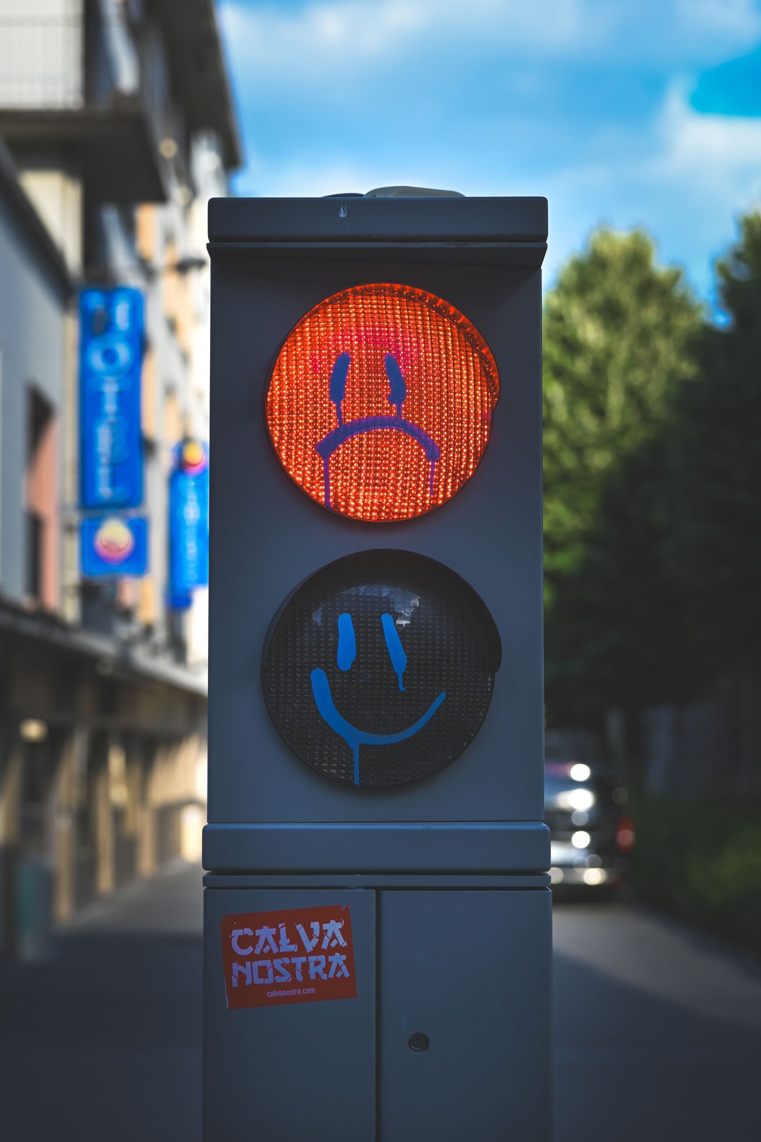 Red pedestrian traffic light with blue smiley and sad face graffiti, street scene in background.