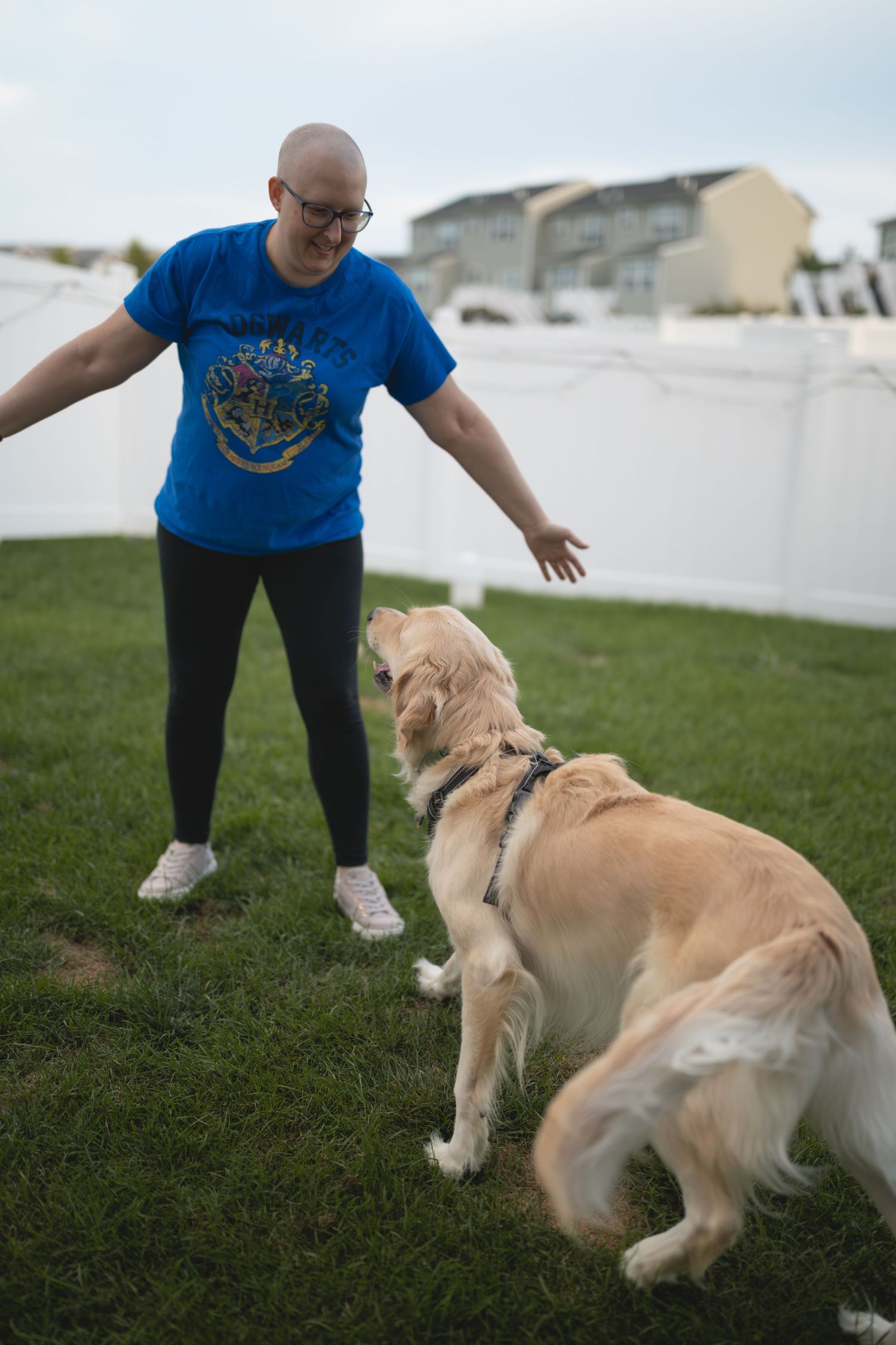 Bald woman in blue Harry Potter shirt playing with golden retriever dog in fenced backyard.