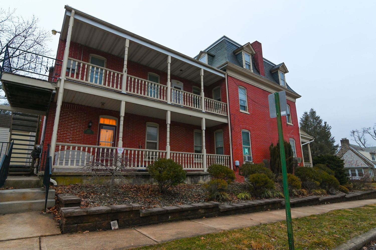 Large two-story red brick house with wrap-around porches, white railings, and dormers on a cloudy day.