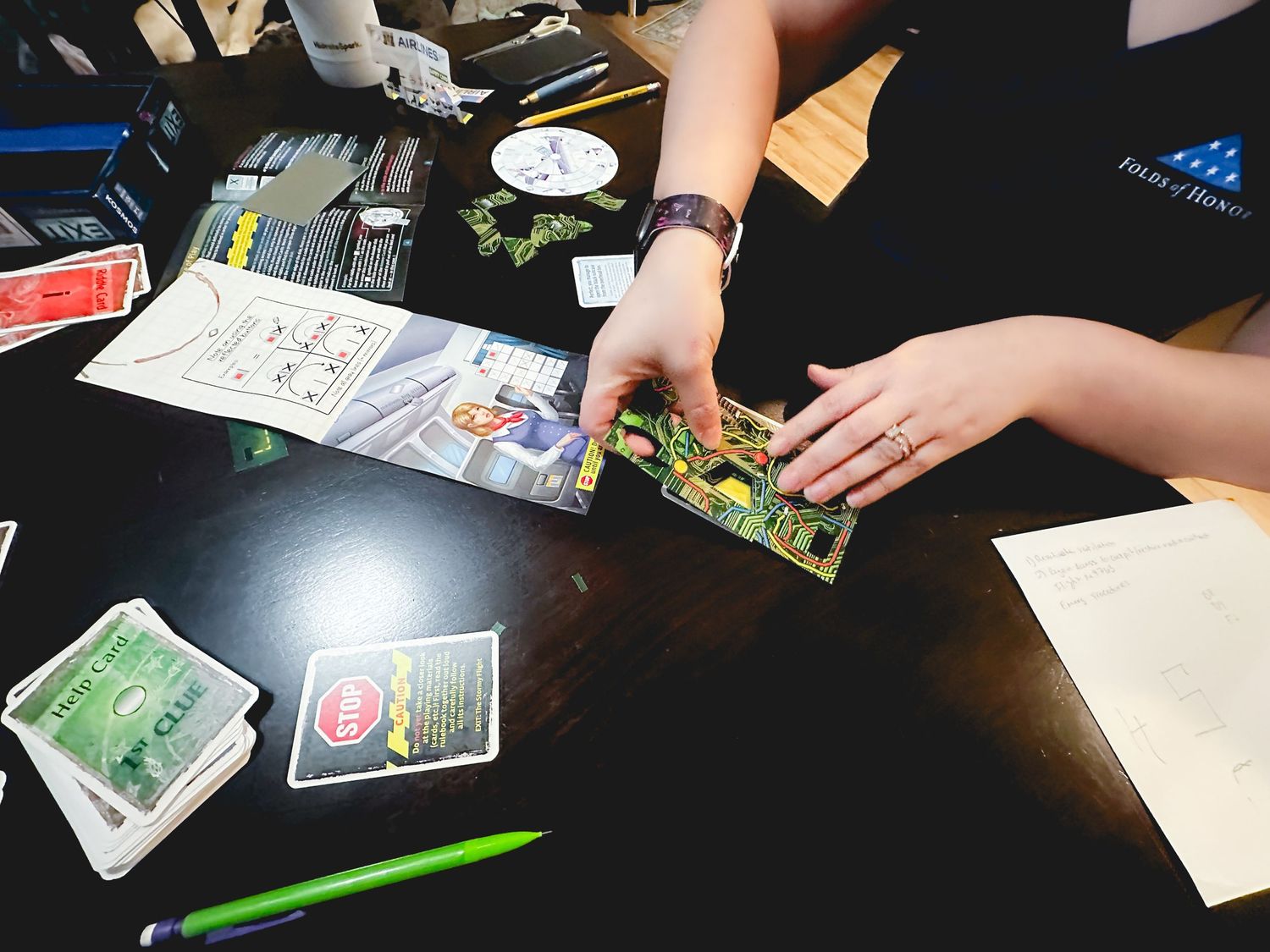 Person holding a colorful board game card over a dark table with game pieces, cards, and a green pen.
