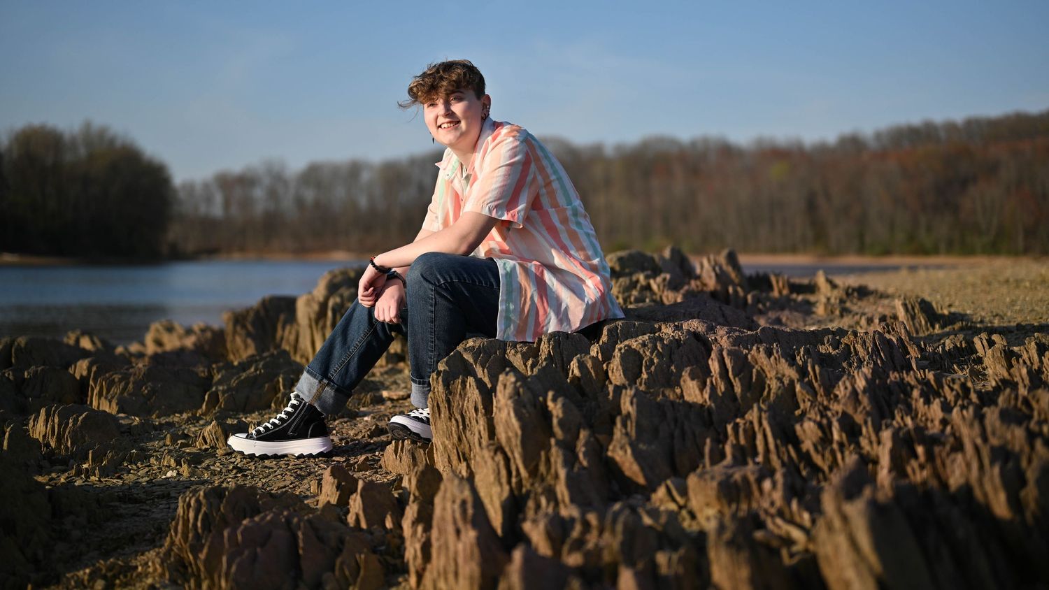 A person sits thoughtfully on rocky terrain near a lake during golden hour against a wooded backdrop.