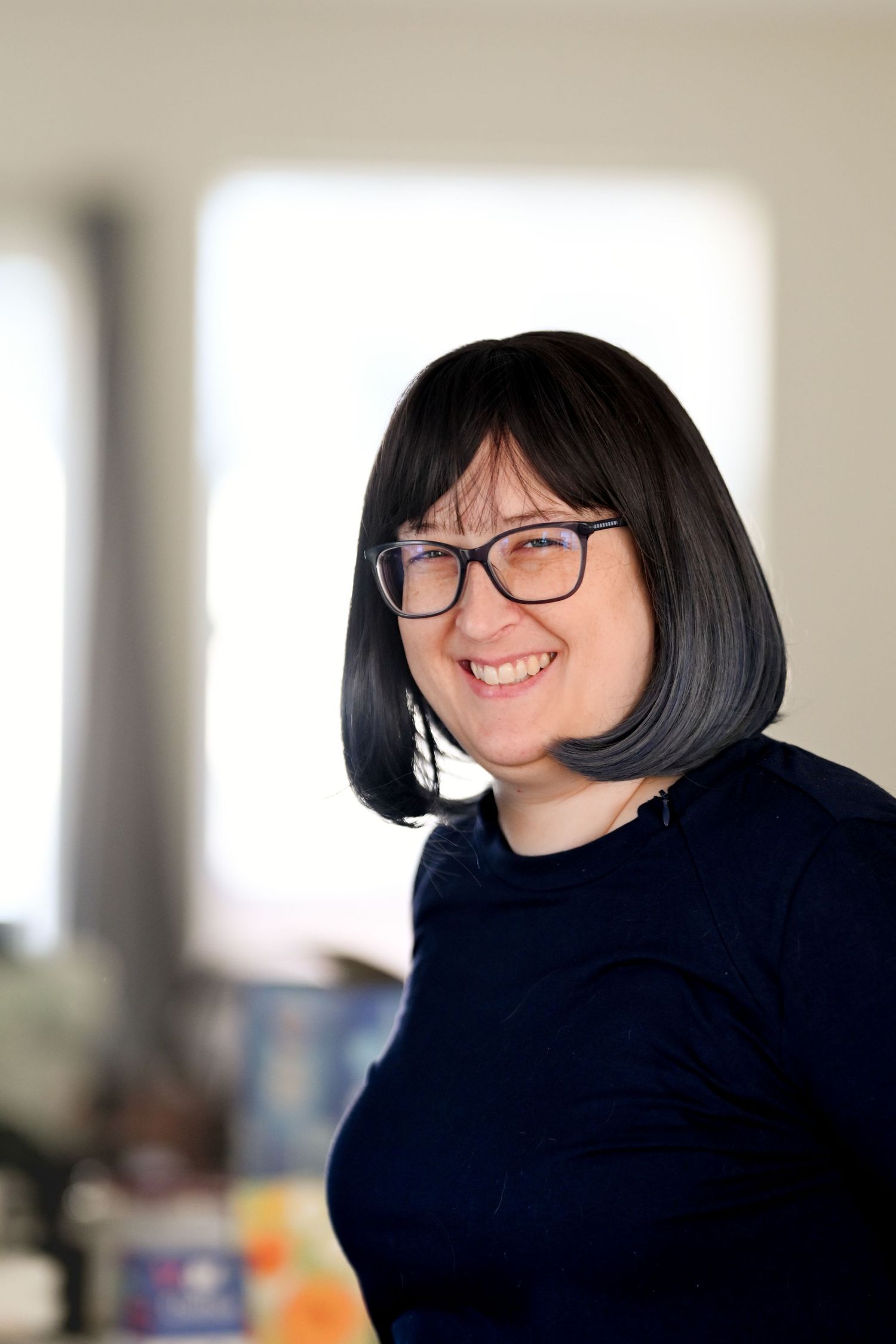 Smiling woman with black bob haircut and glasses wearing a navy blue top in a bright indoor setting.