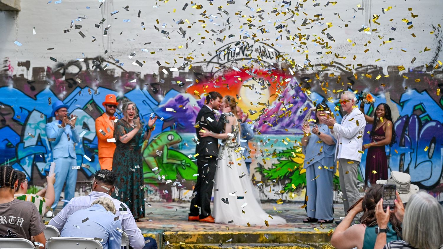 Wedding couple celebrates under shower of confetti with colorful graffiti wall backdrop as guests cheer in urban setting.