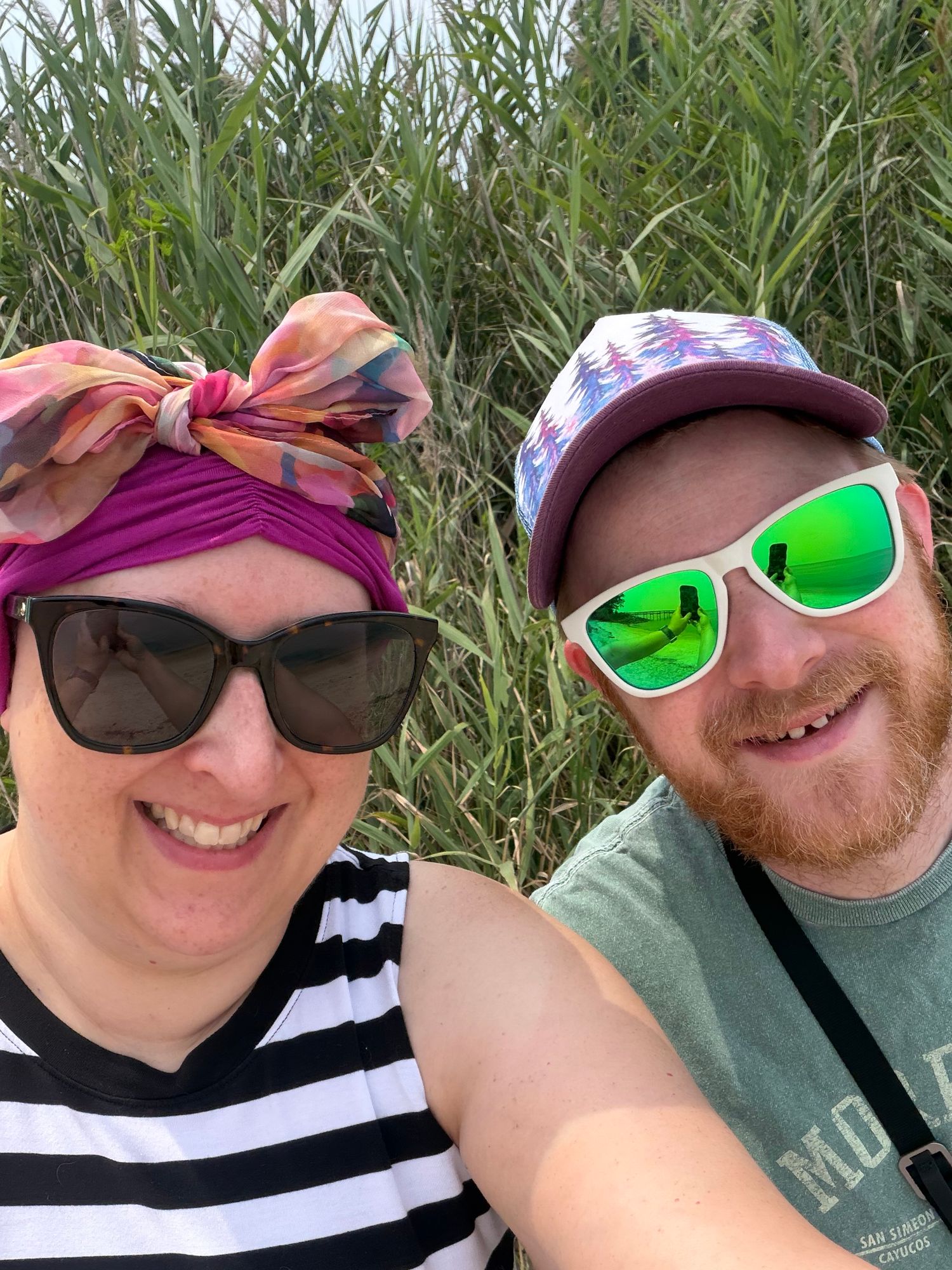 Two smiling people wearing sunglasses and hats take a selfie outdoors near tall green reeds.