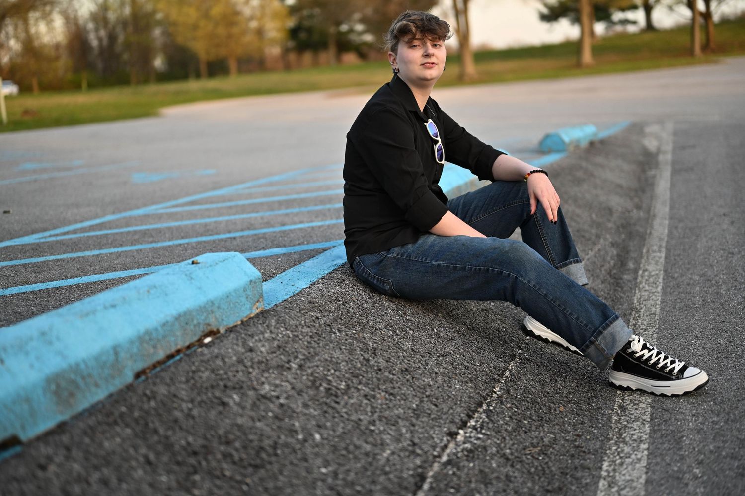 Someone in casual wear sits on a blue-painted curb in an empty parking lot wearing sunglasses and sneakers.