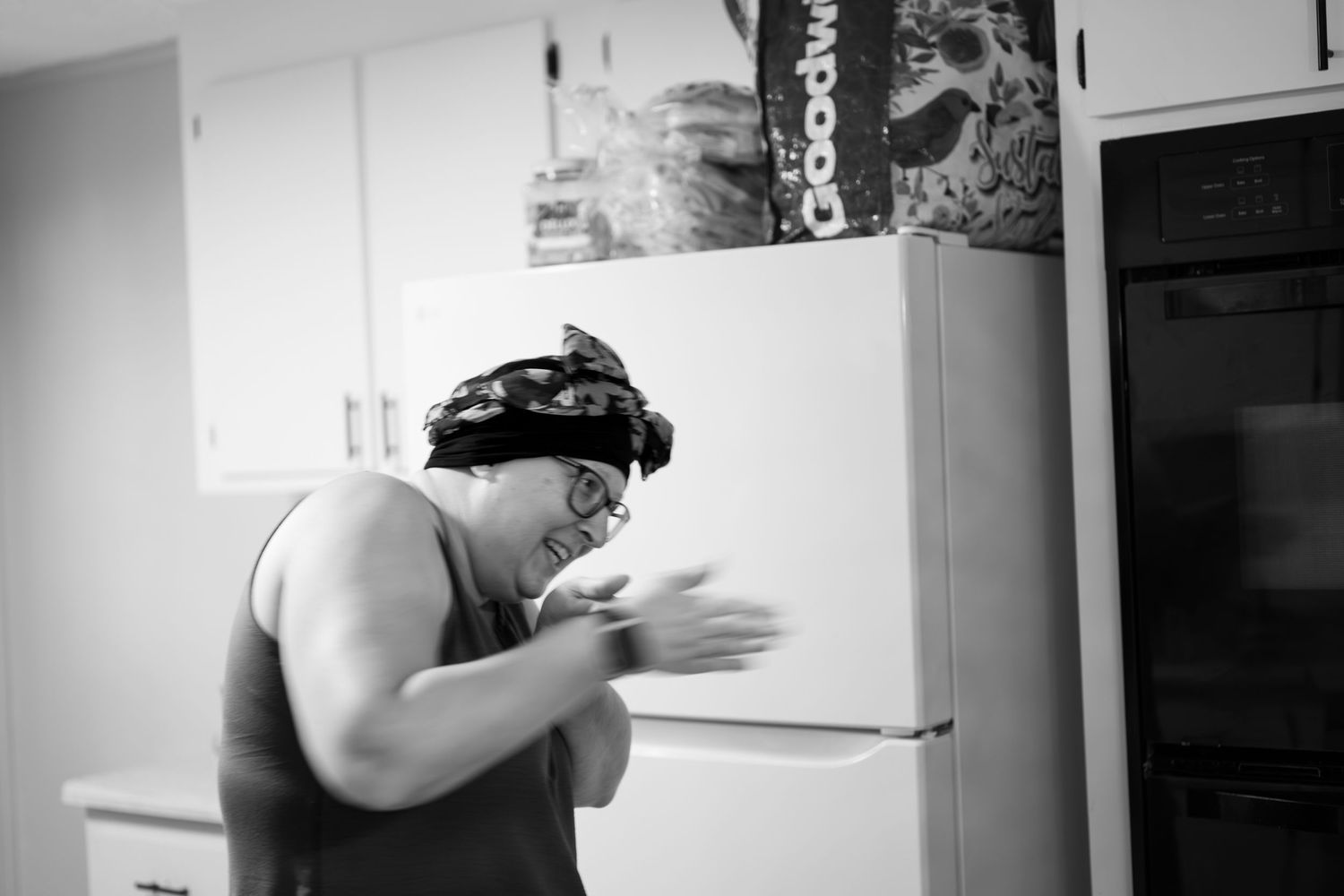Man in bandana and tank top clapping hands near refrigerator in kitchen, black and white photo.