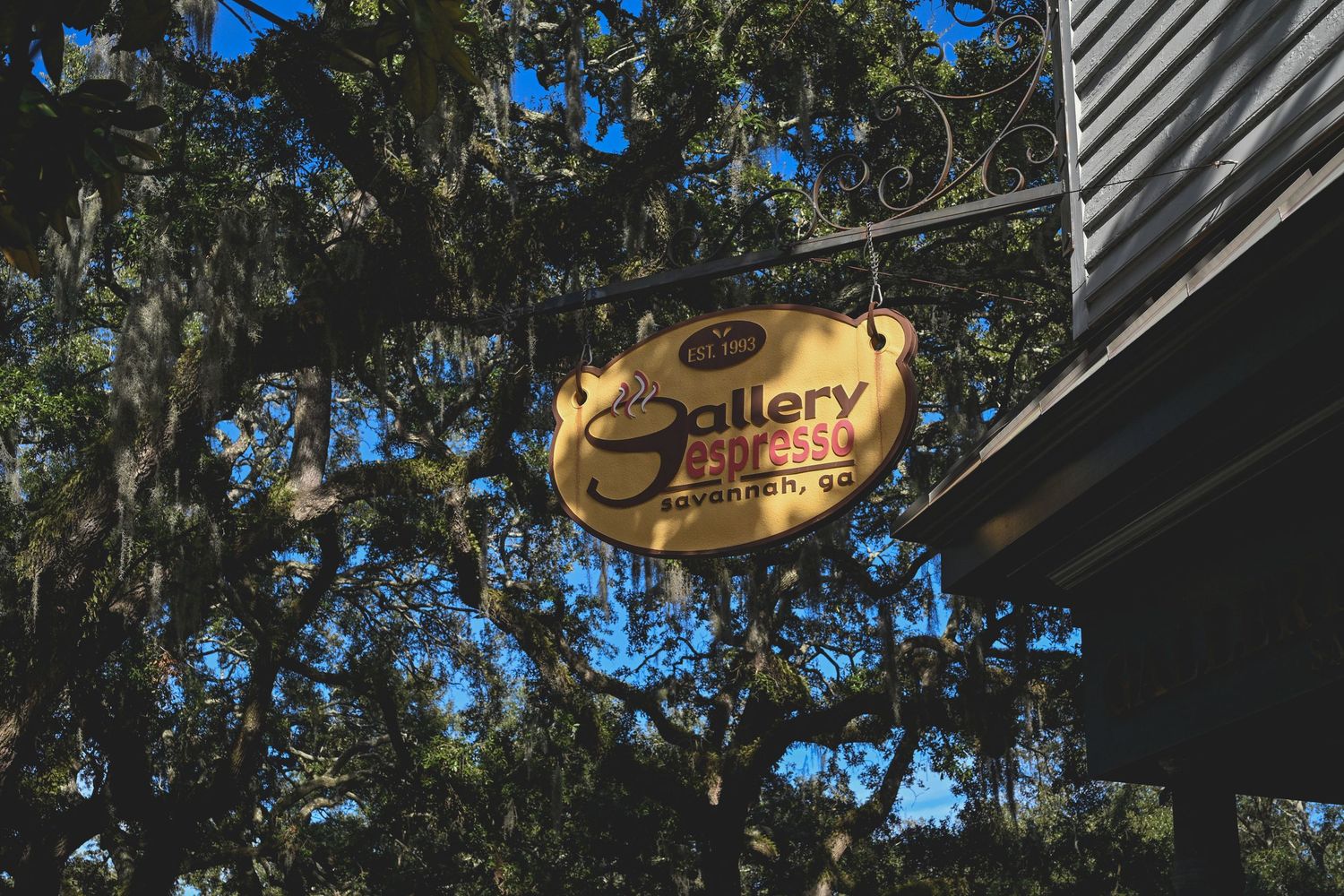 A series of gold-colored business signs hang from a dark wooden building exterior against a blue sky and green trees.