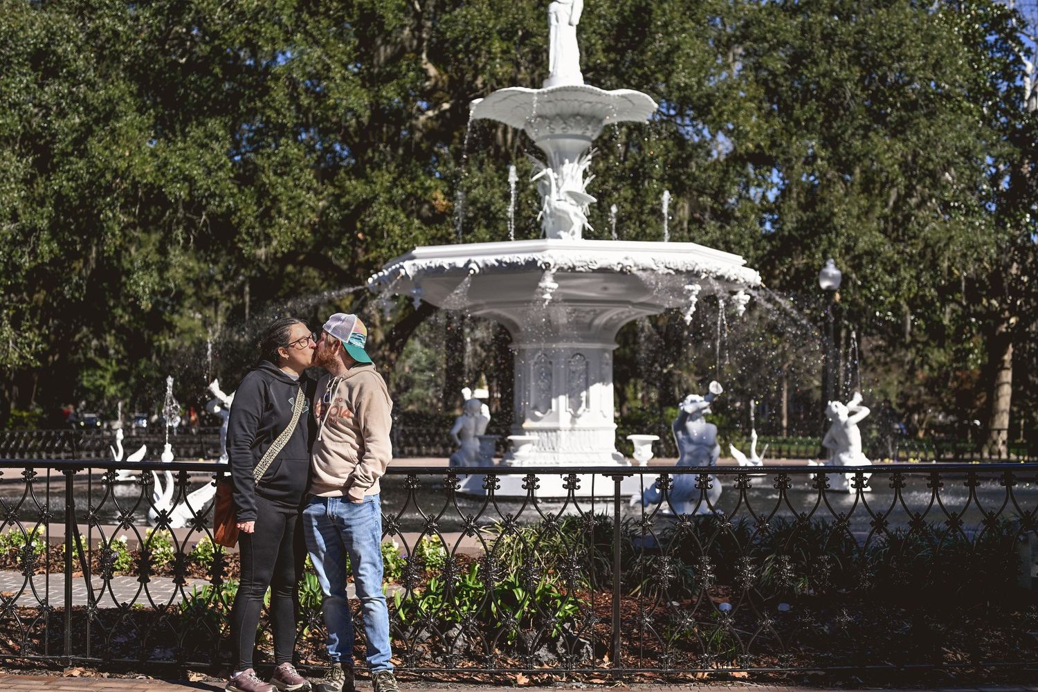 Panoramic view of a park fountain surrounded by lush greenery and people enjoying a sunny day.