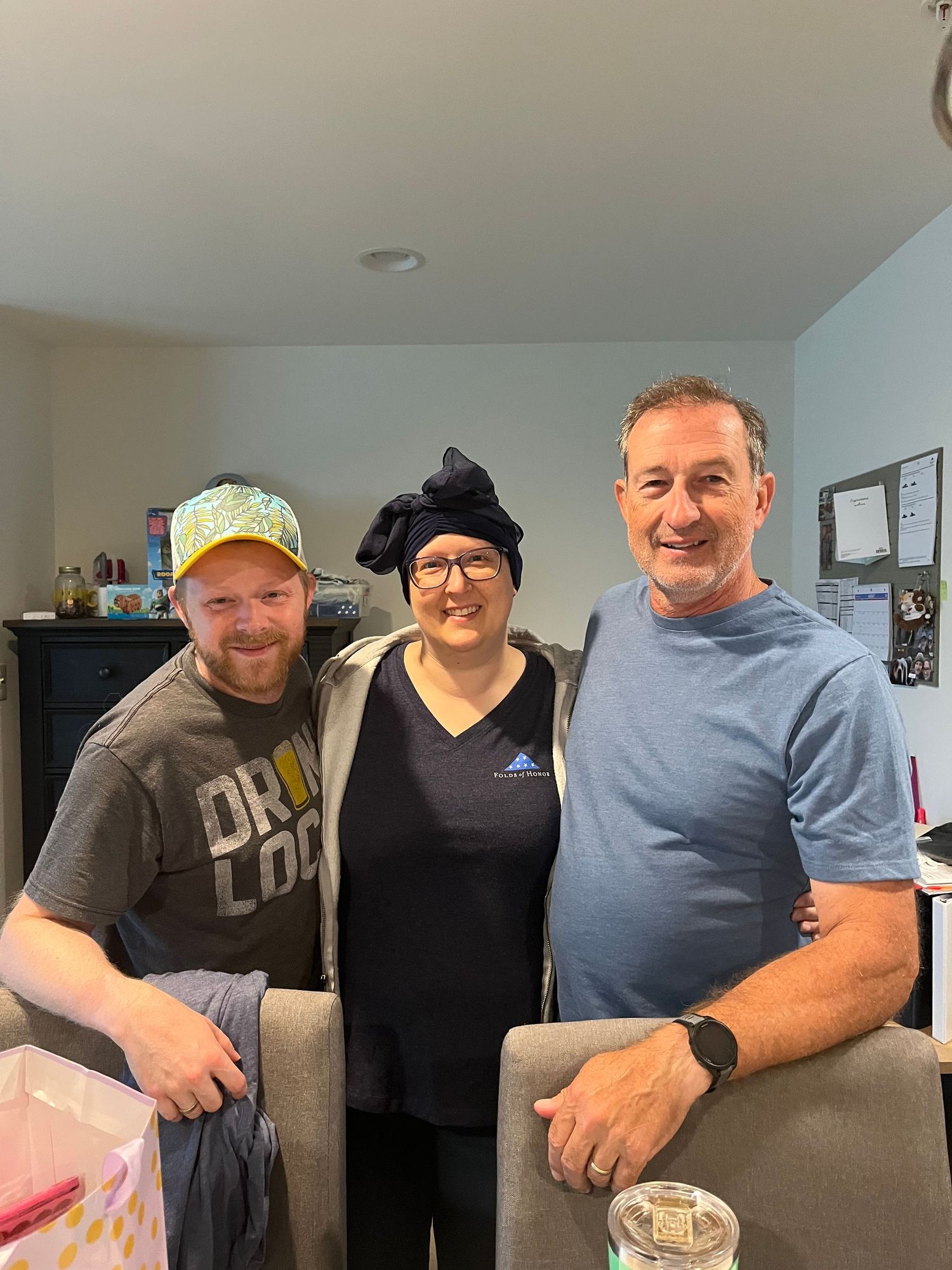 Three people smiling together indoors, two men and a woman wearing a black head wrap, posing for a casual group photo.
