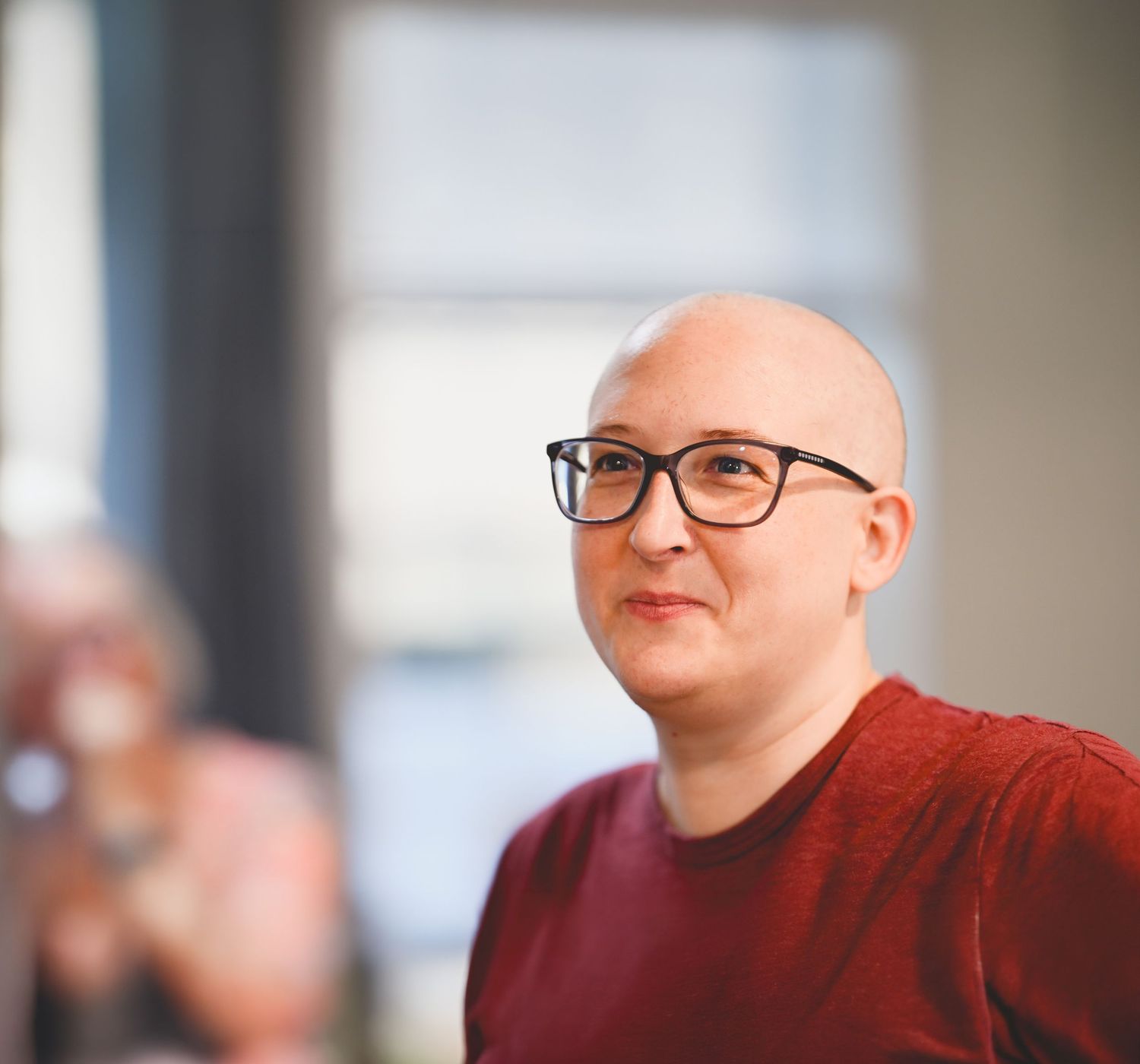 Bald person wearing glasses and a red shirt, sitting indoors with a blurred background.