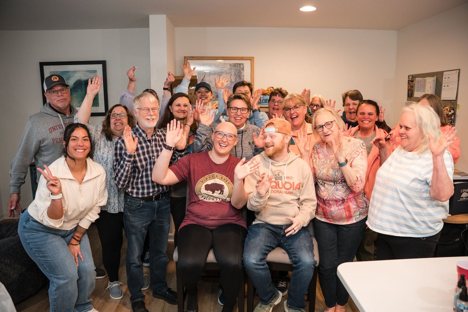 Large group of cheerful adults celebrating together indoors, smiling and raising hands in a cozy living room setting.