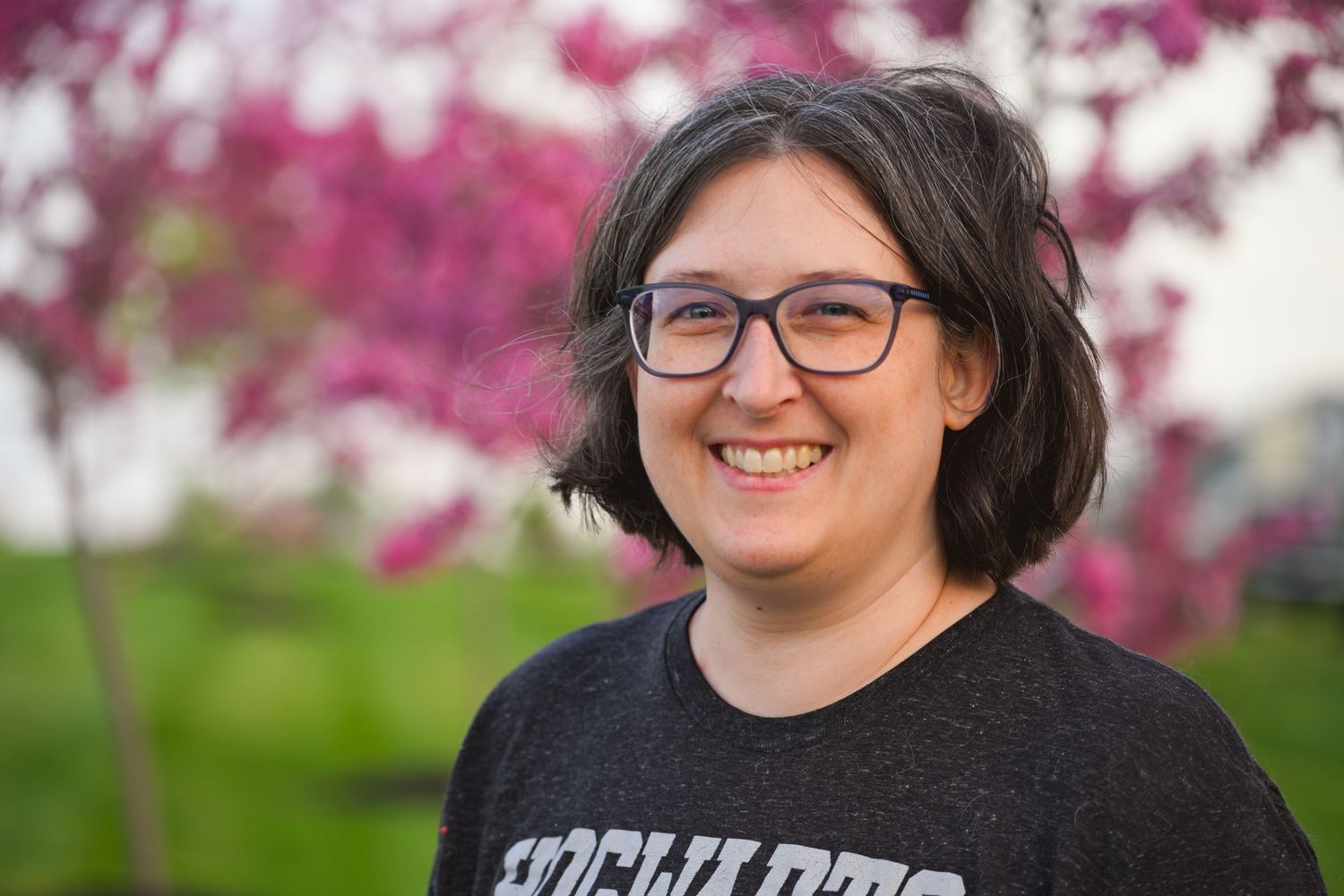 Smiling woman with glasses and dark hair wearing a Hogwarts shirt, standing outdoors near pink flowering trees.