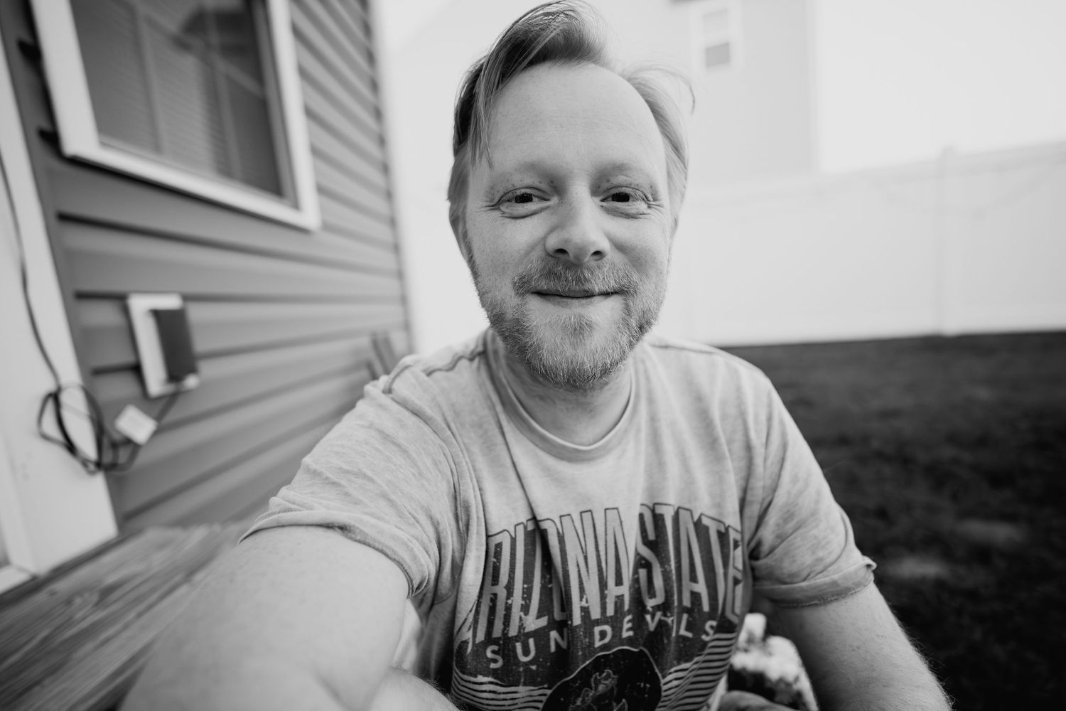 Black and white selfie of a smiling bearded man in an Arizona State Sun Devils t-shirt outdoors.