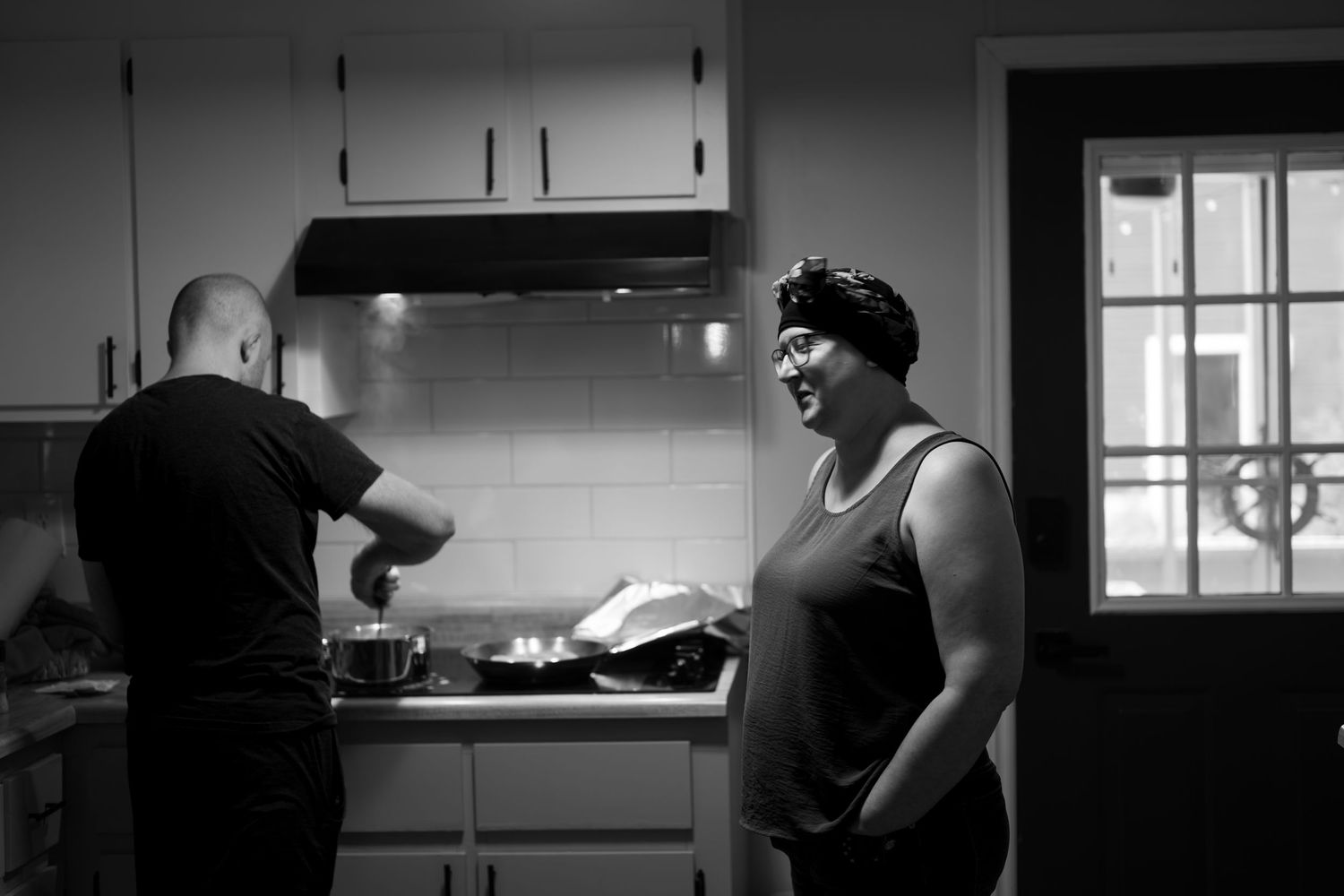 Black and white photo of two people in a kitchen, one cooking at the stove while the other laughs nearby.