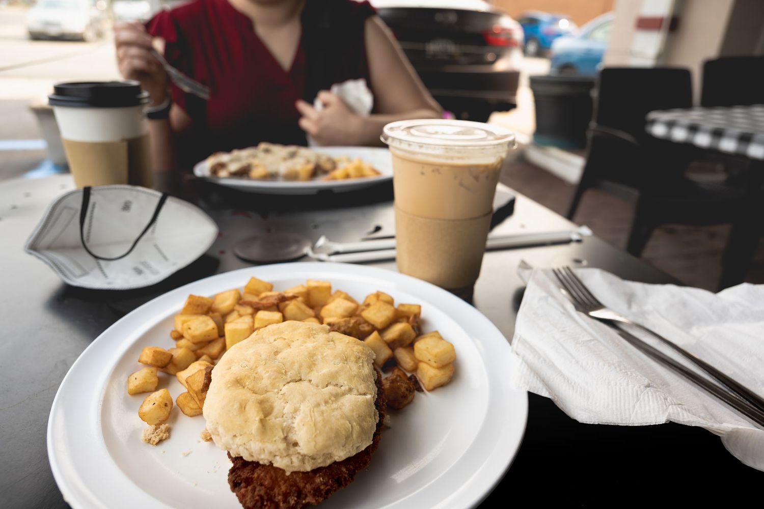 A fried chicken biscuit sandwich with crispy potatoes on a white plate, beside an iced latte at a casual breakfast restaurant.