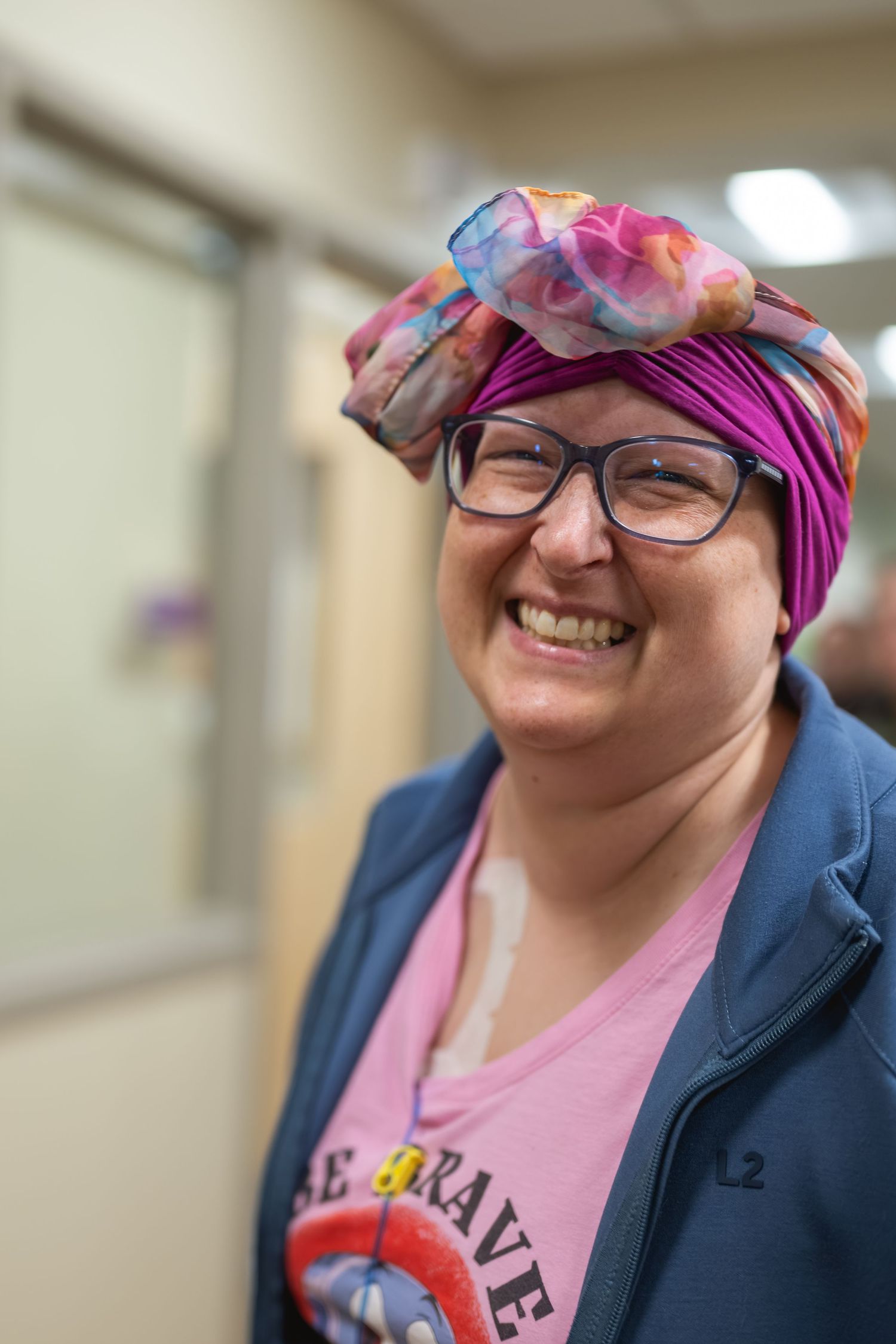 Smiling woman wearing colorful head wrap and glasses in a hallway, pink shirt reading brave.