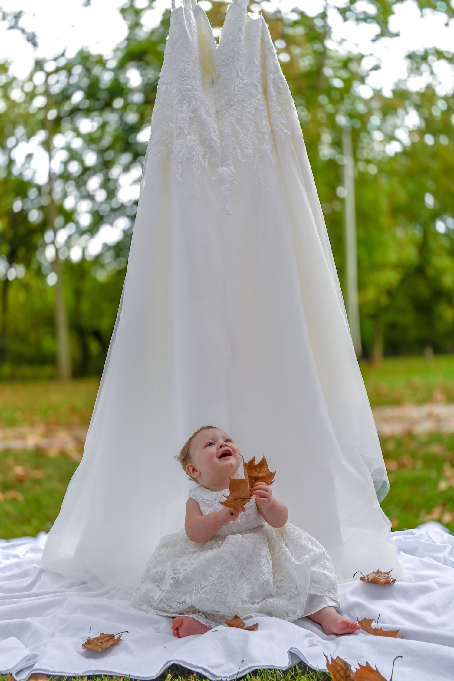 A small baby in white dress sits under a hanging wedding gown outdoors while playing with autumn leaves on a blanket.