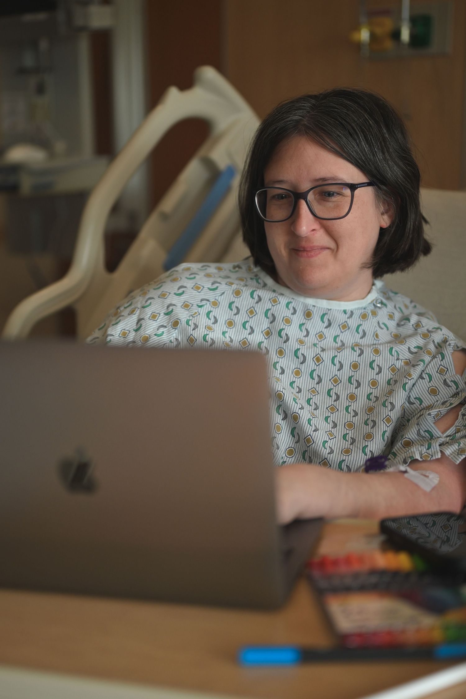 Woman in hospital gown working on MacBook laptop at bedside table with colored markers nearby.