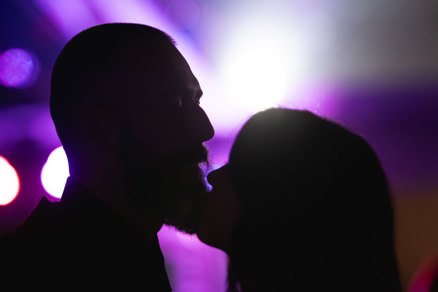Silhouette of a couple about to kiss in profile against glowing purple and pink stage lights at a nightclub or party.